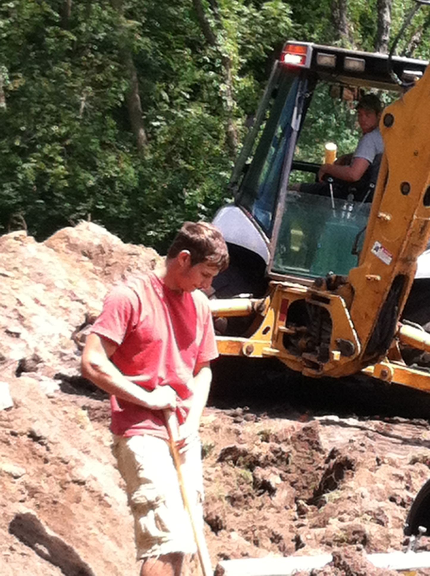A person in a red shirt and tan shorts works with a shovel near a yellow backhoe in a dirt-filled outdoor construction area.