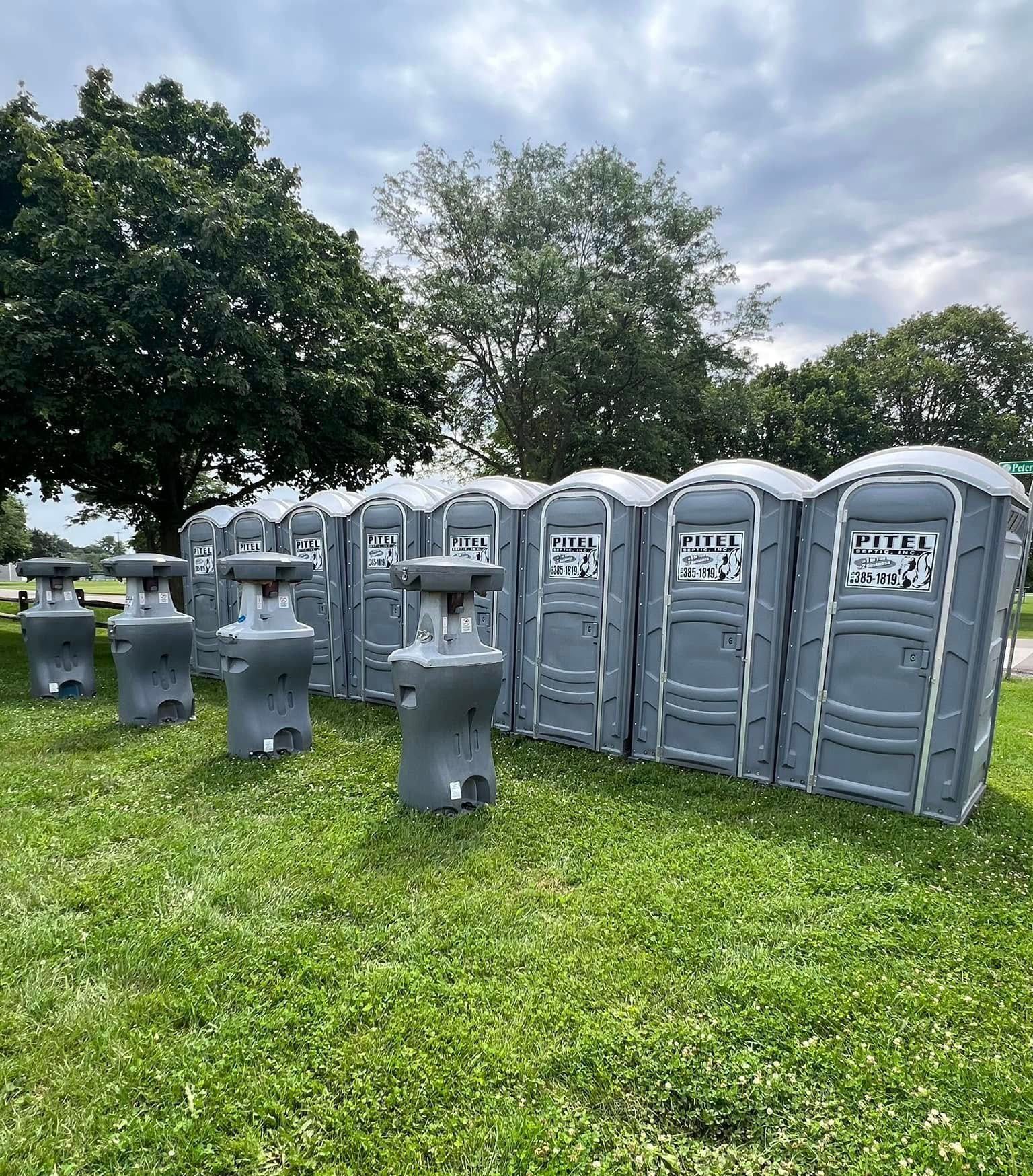 Portable toilets and handwashing stations on grassy field. Gray, outdoor restroom facilities. Cloudy sky, trees.