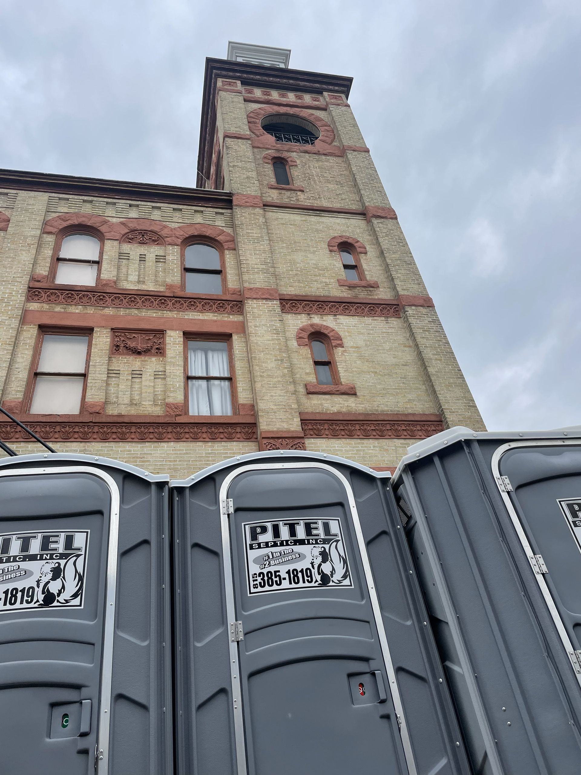 A tall brick building with a clock tower and several portable toilets in front of it