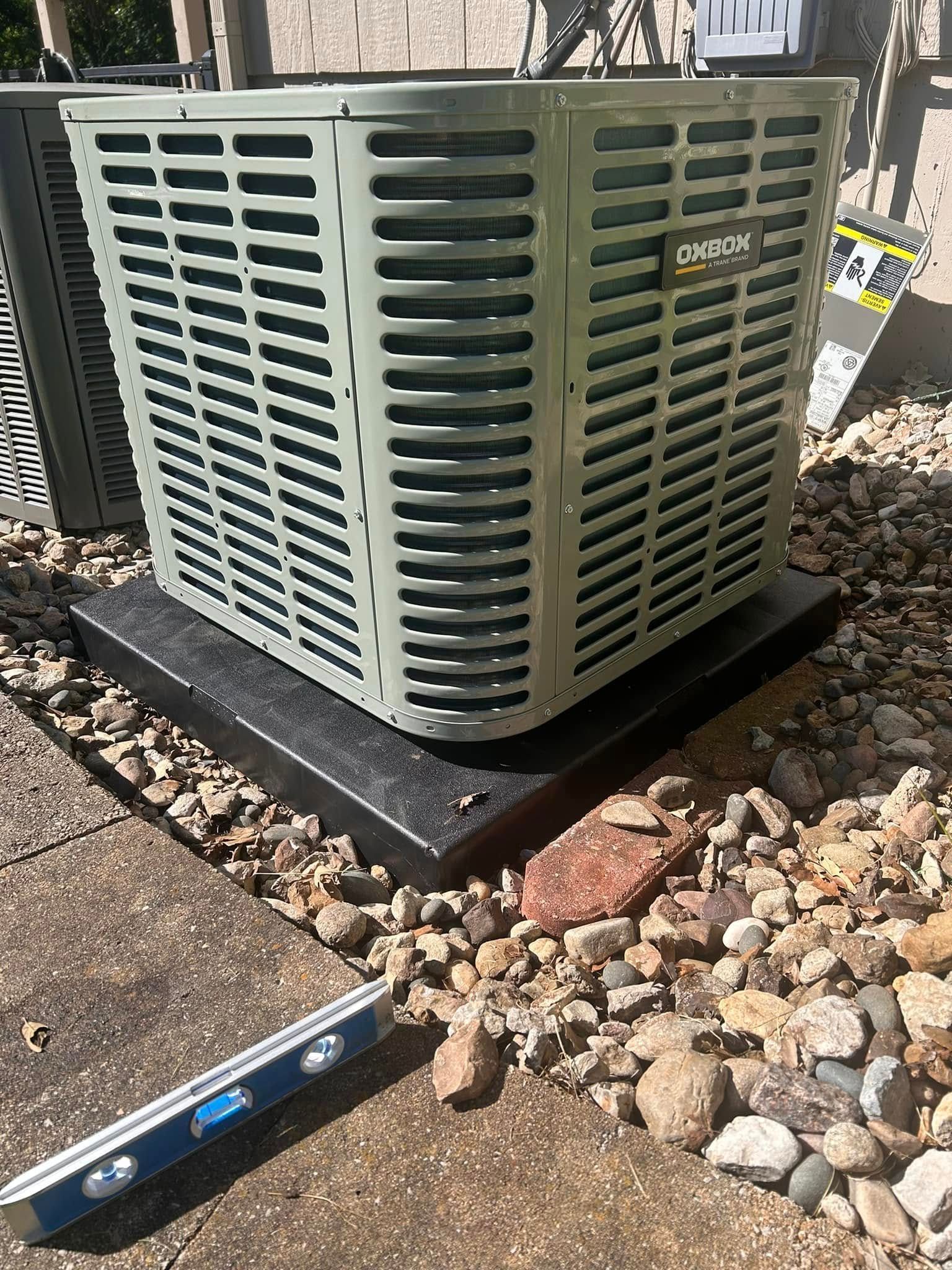 AC unit on a black platform, surrounded by rocks, next to a paved area; a level is at the bottom left.