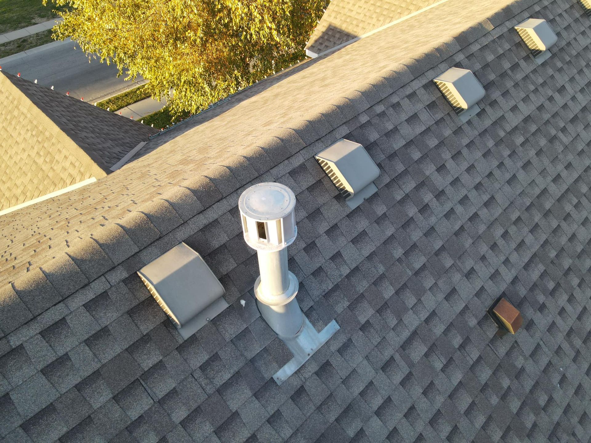 Overhead view of a shingled roof with several vents, including a tall, round one.