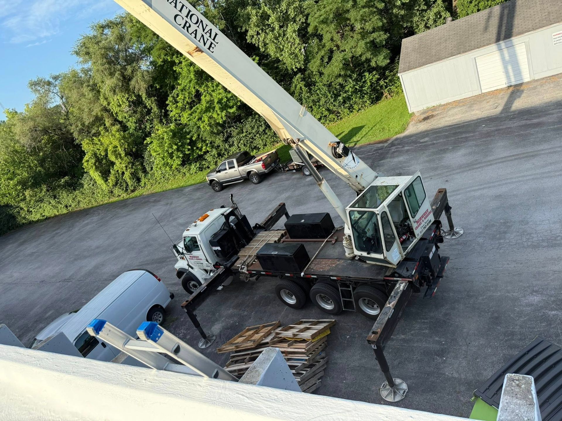 Crane truck lifting in a parking lot near green trees and a white building.