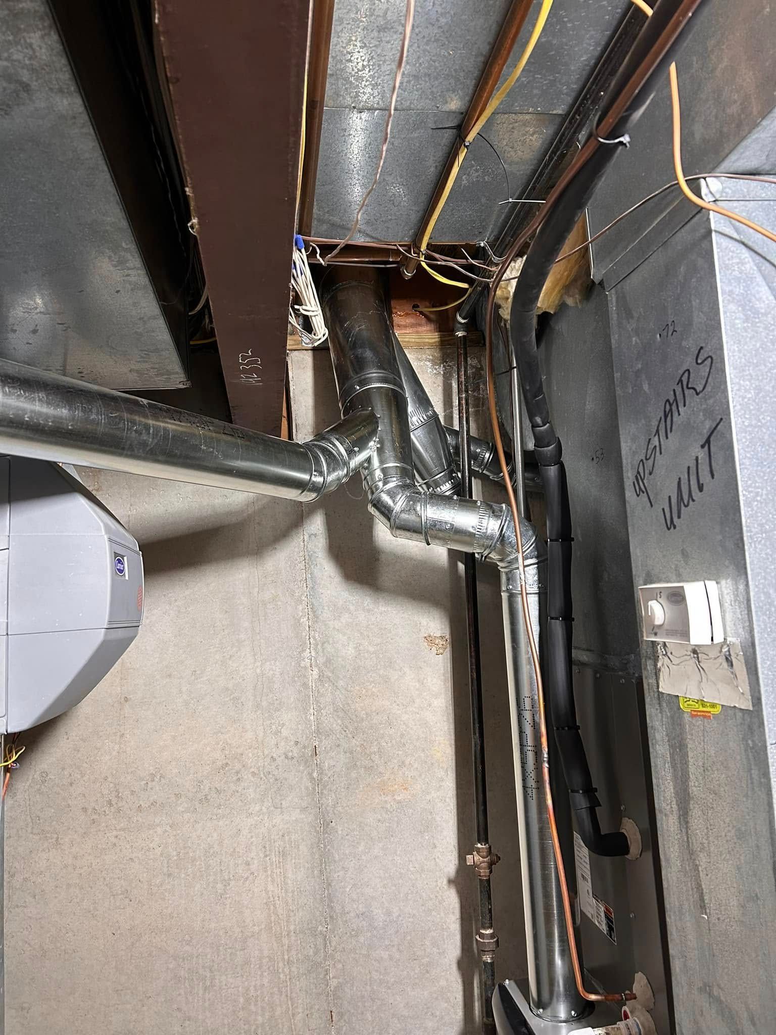 Metal pipes and wires near a concrete wall. Grey and black insulation, some overhead brown beams.