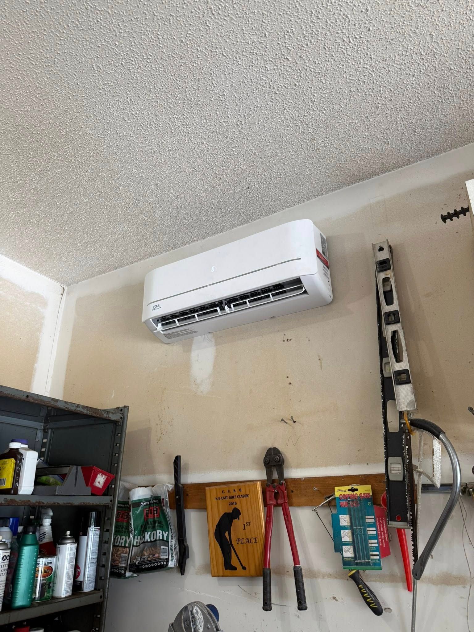 An air conditioner is mounted on a garage wall, with tools and a shelf below.