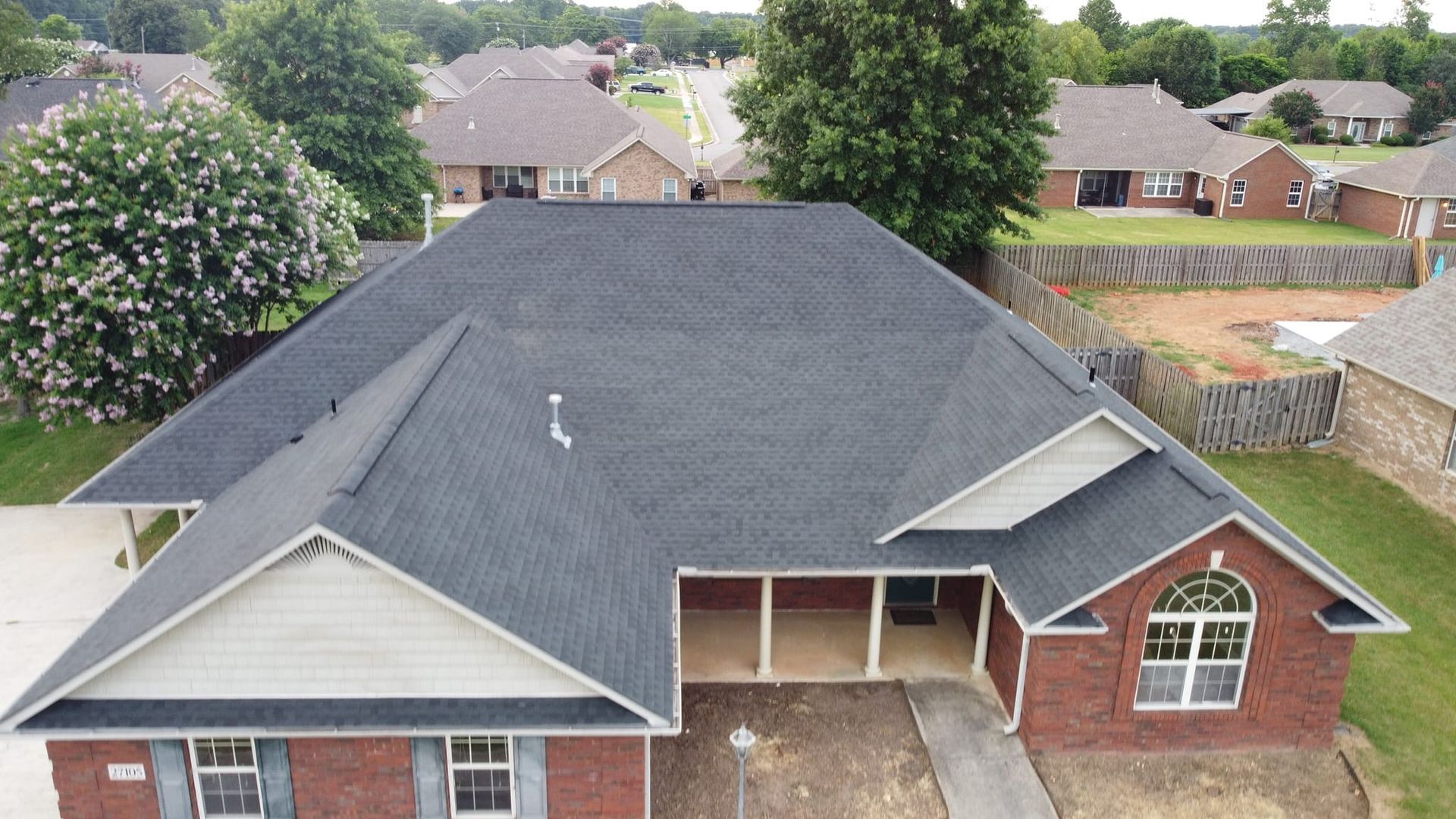 A front-facing aerial view shows a residential home with a newly installed dark shingle roof in a suburban neighborhood.