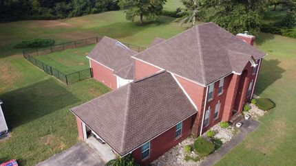An aerial, high-angle view shows a two-story red brick house with a brown shingled roof, featuring a fenced-in side yard and a large green lawn bordered by trees.