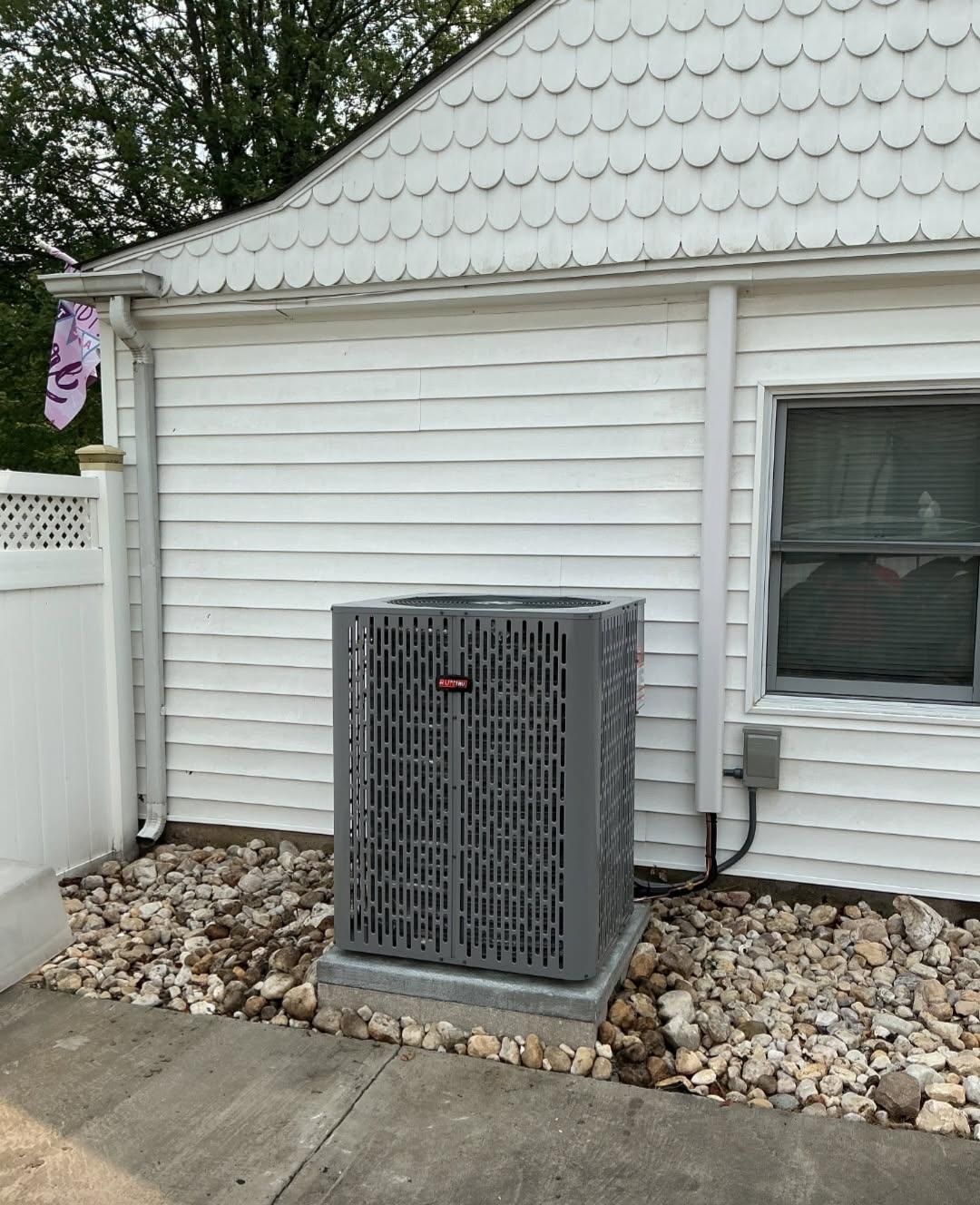 A gray air conditioning unit sits on a concrete base next to a white house with a rock bed.