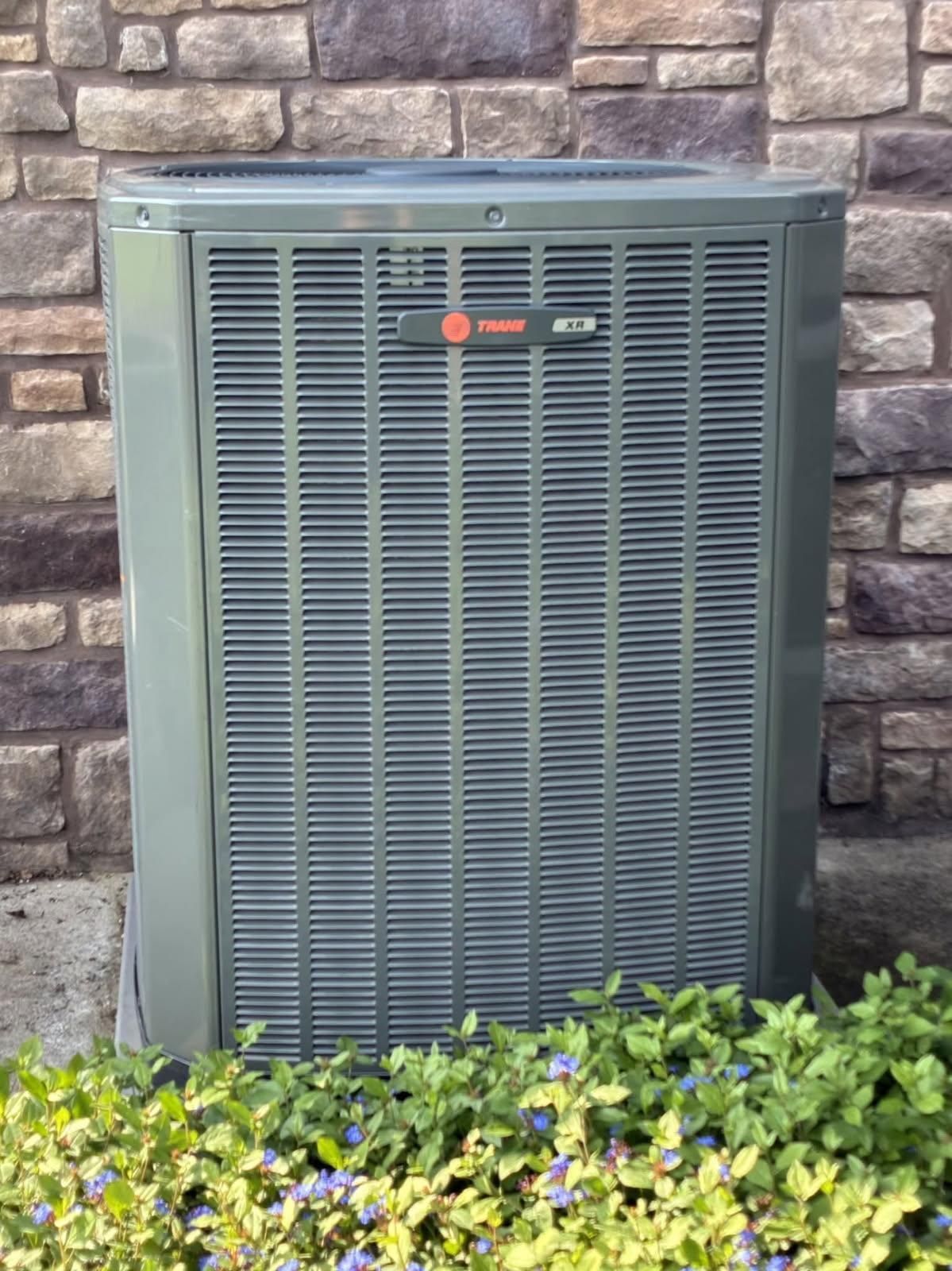 Gray Trane air conditioner unit outside, fronted by green bushes, against a brick wall.