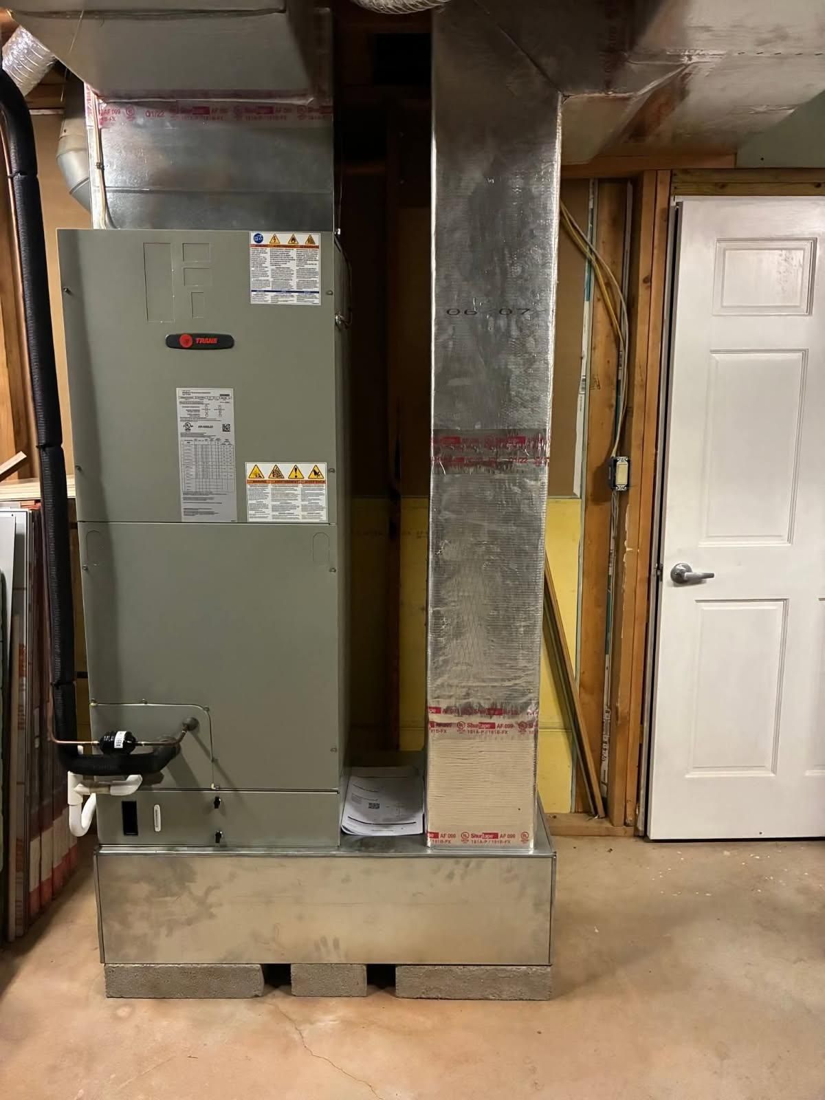 A gray HVAC unit is between ductwork and a white door in a basement.