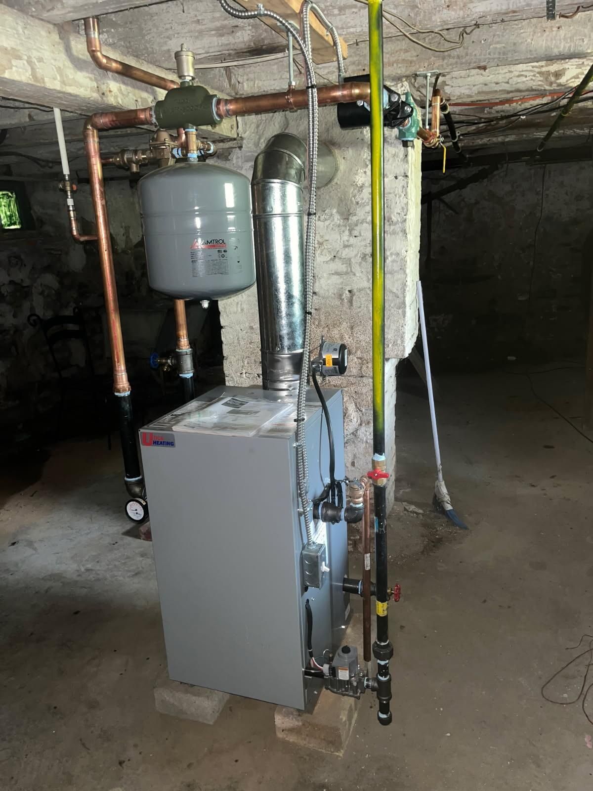 A gray furnace with a chimney in a concrete basement, with copper piping and an expansion tank.