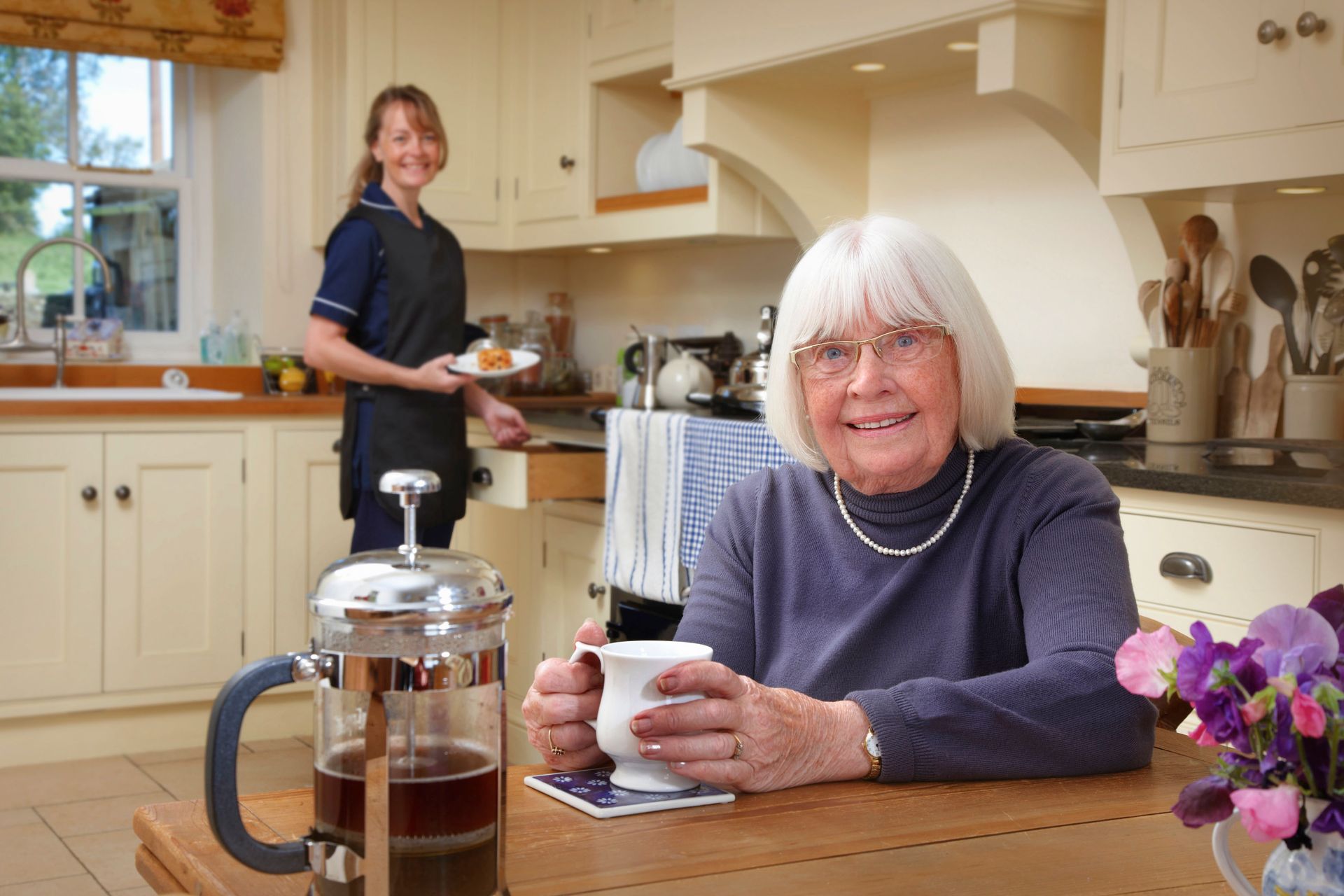 An elderly woman is sitting at a table in a kitchen holding a cup of coffee.