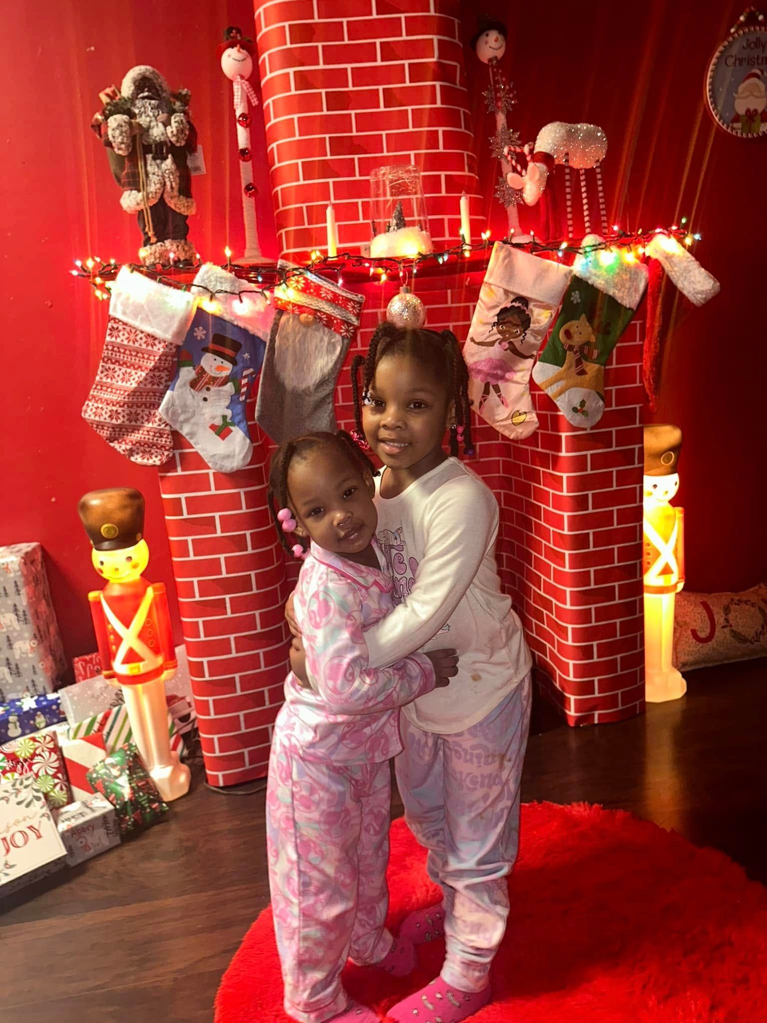 Two young girls in pajamas hugging in front of a Christmas-themed fireplace with stockings and presents.