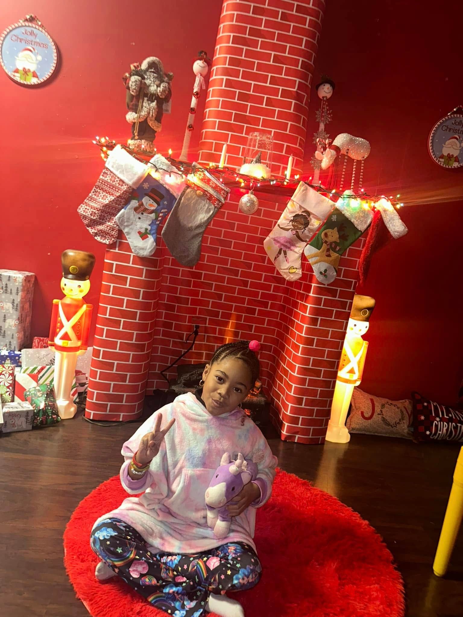 Girl in pajamas sits on red rug in front of a Christmas fireplace.