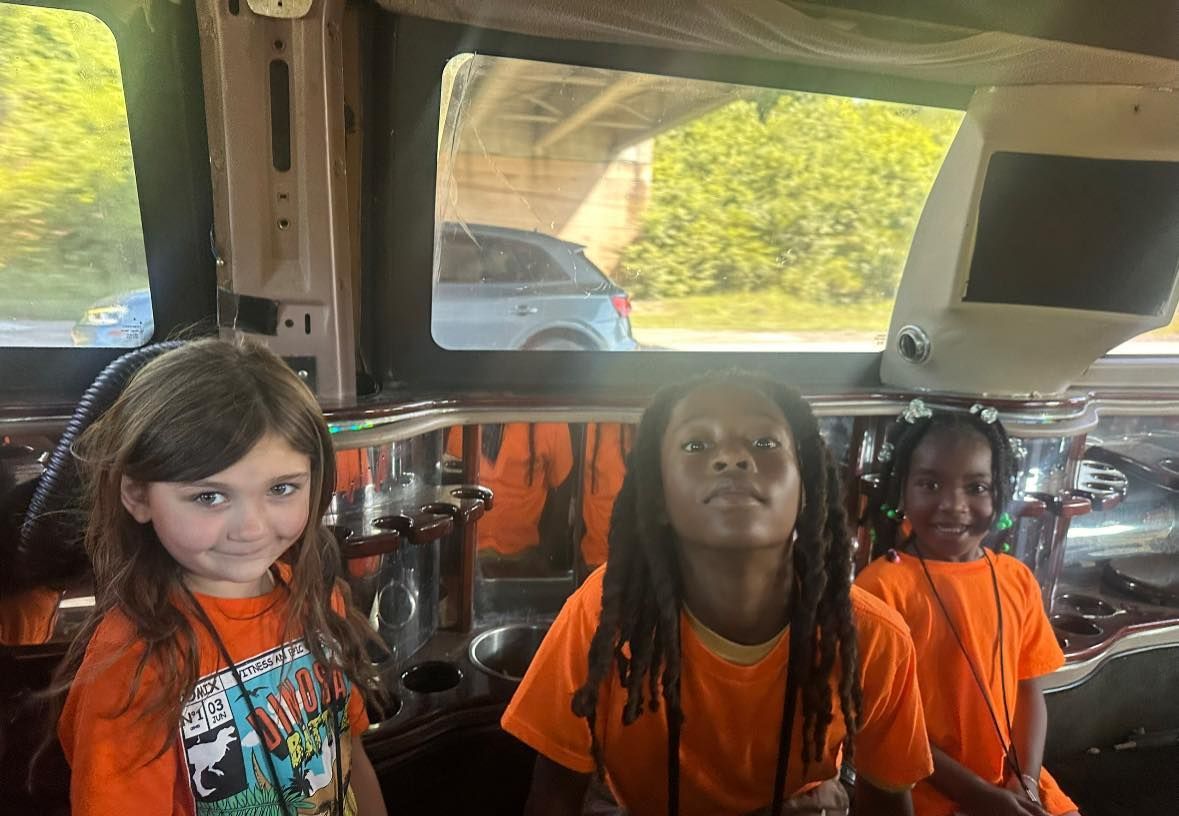 Three children in orange shirts inside a vehicle, looking at the camera.