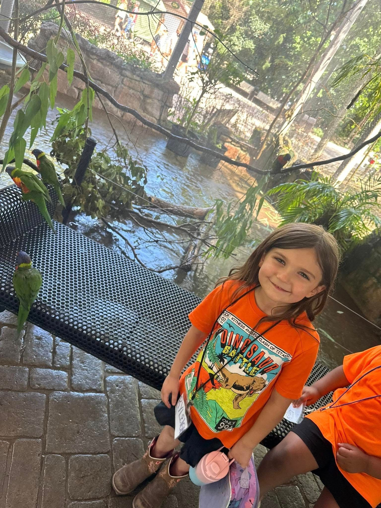 Girl in orange shirt smiles at the camera by a bird enclosure, with green parrots inside.