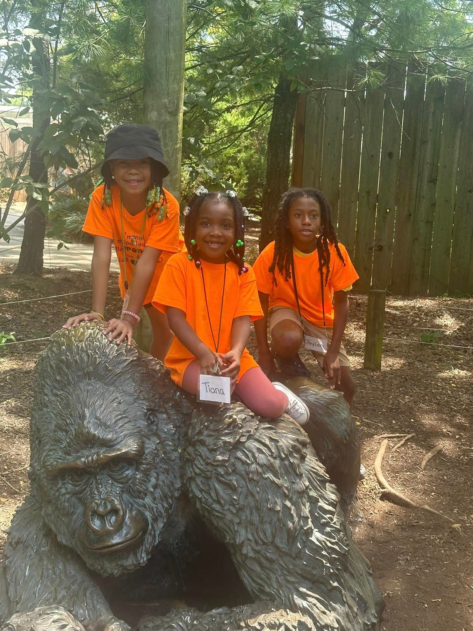 Three smiling children in orange shirts pose on a gorilla statue in a park.