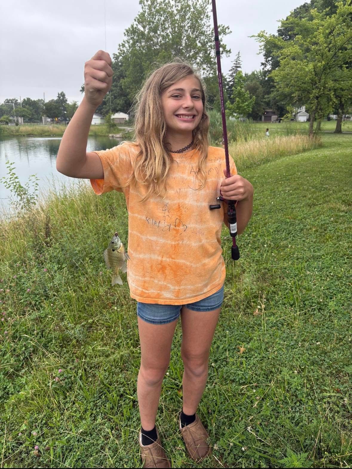 Girl in orange shirt holds up a small fish, standing by a pond with a fishing rod.