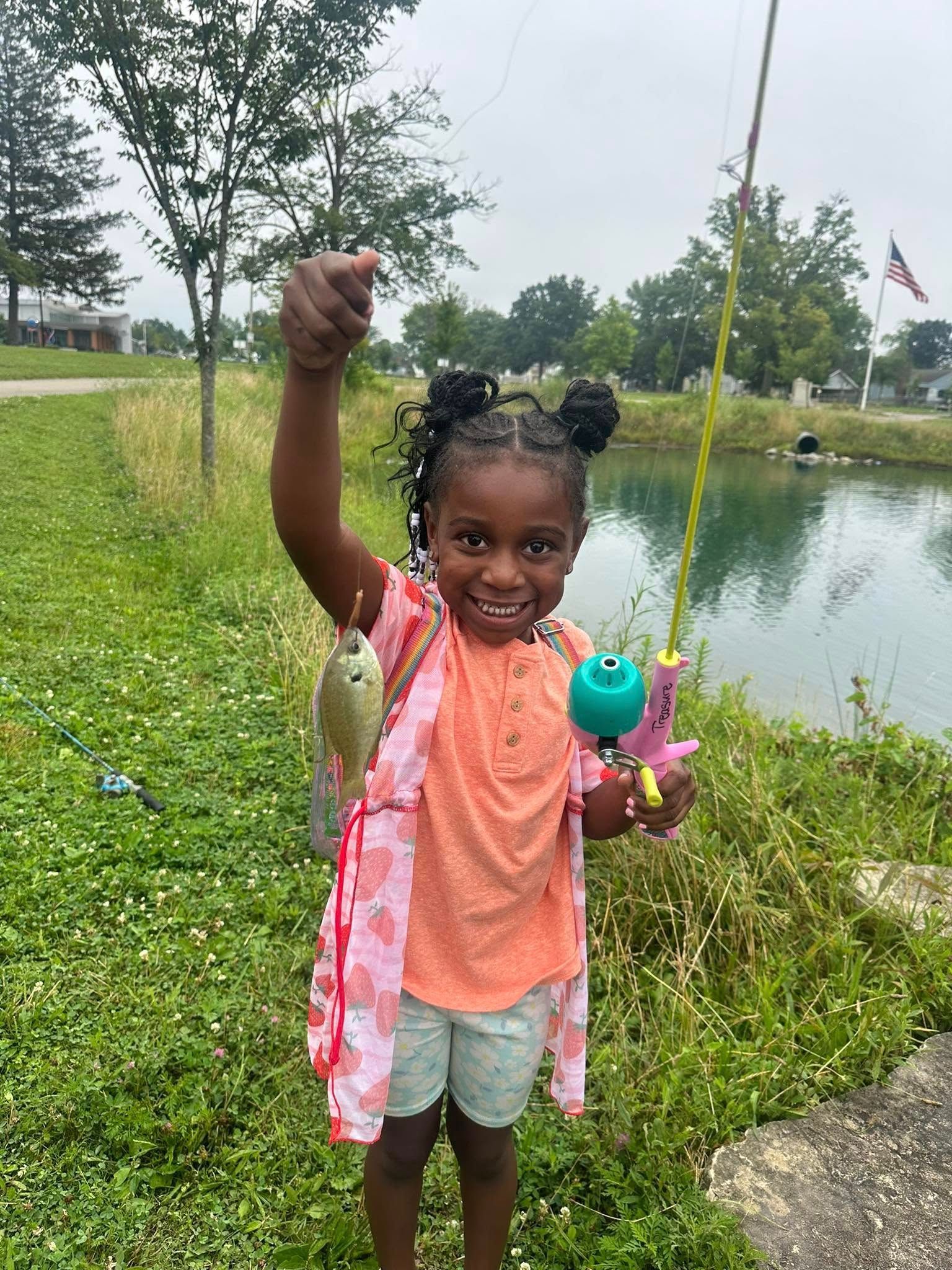 Young girl proudly displays fish she caught, next to a pond with fishing rod.