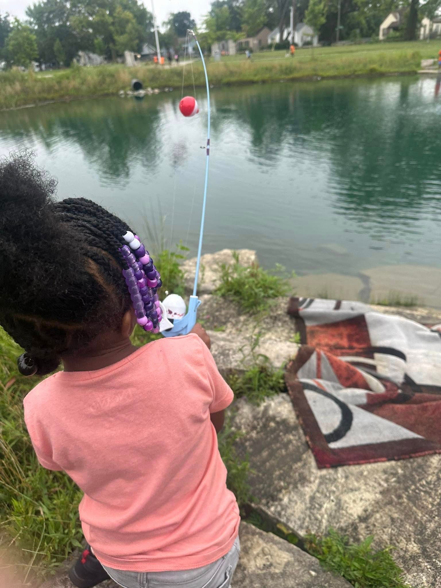 Young child with braids fishes from a bank near water, using a blue pole with a red ball on top.
