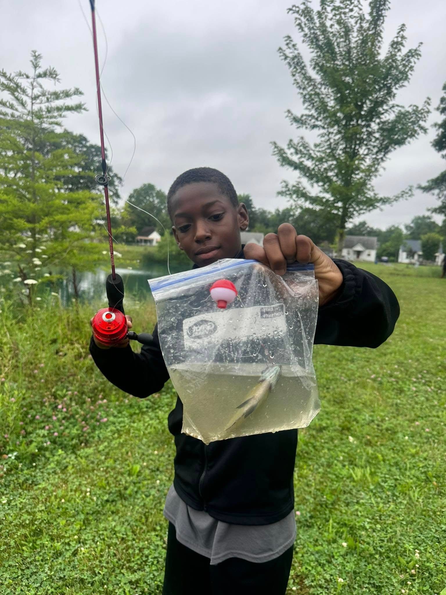Boy holding a bag with a small fish, standing near a pond with a fishing rod.