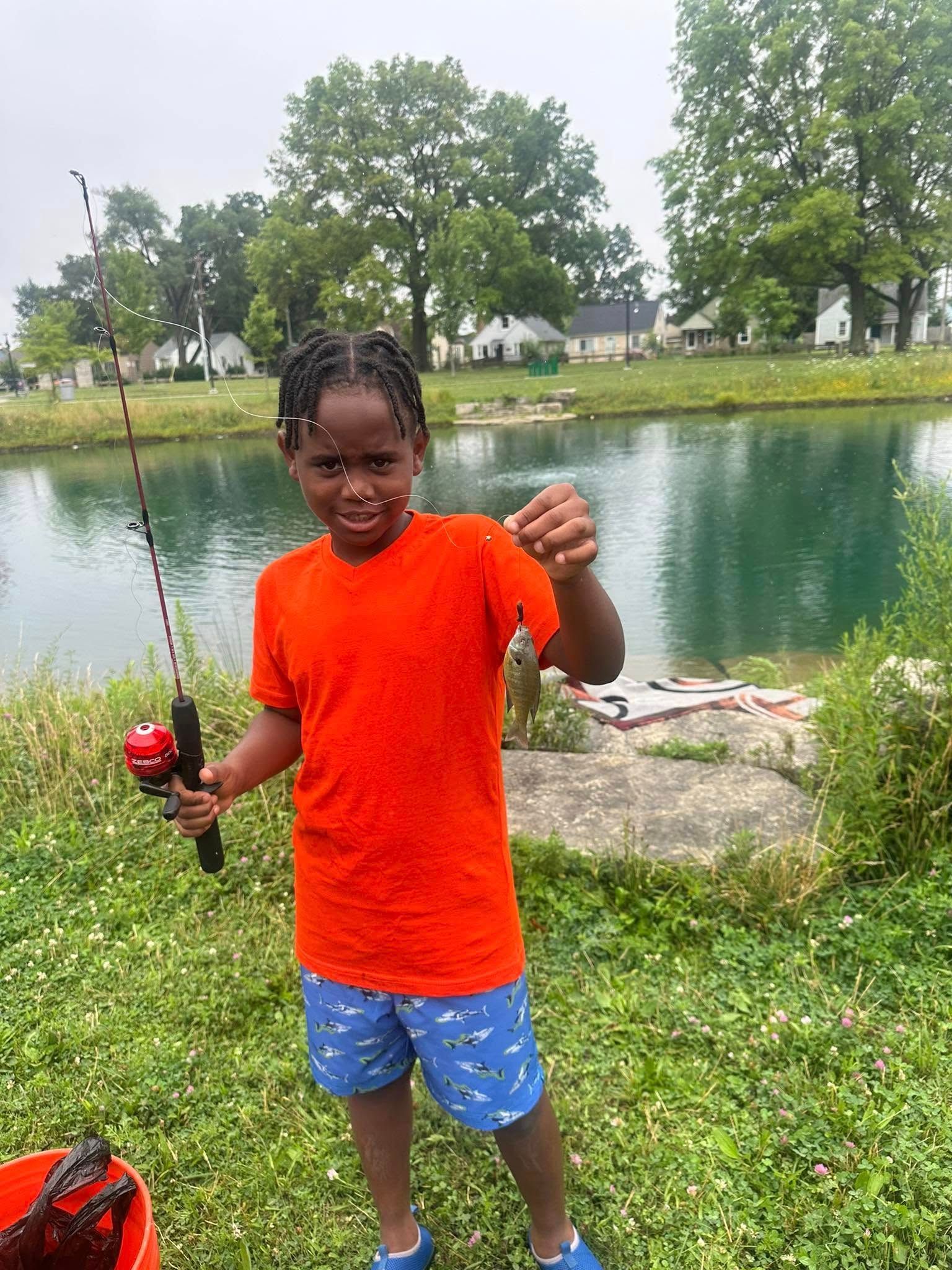 Young boy in orange shirt and blue shorts fishing, holding up a small fish near a pond.