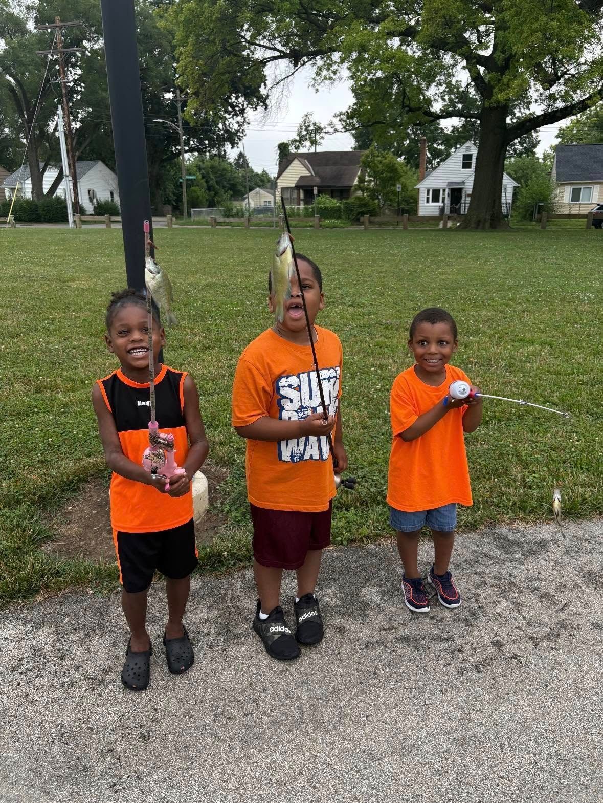Three children proudly display fish they caught while smiling; green grass, gray road, houses in background.
