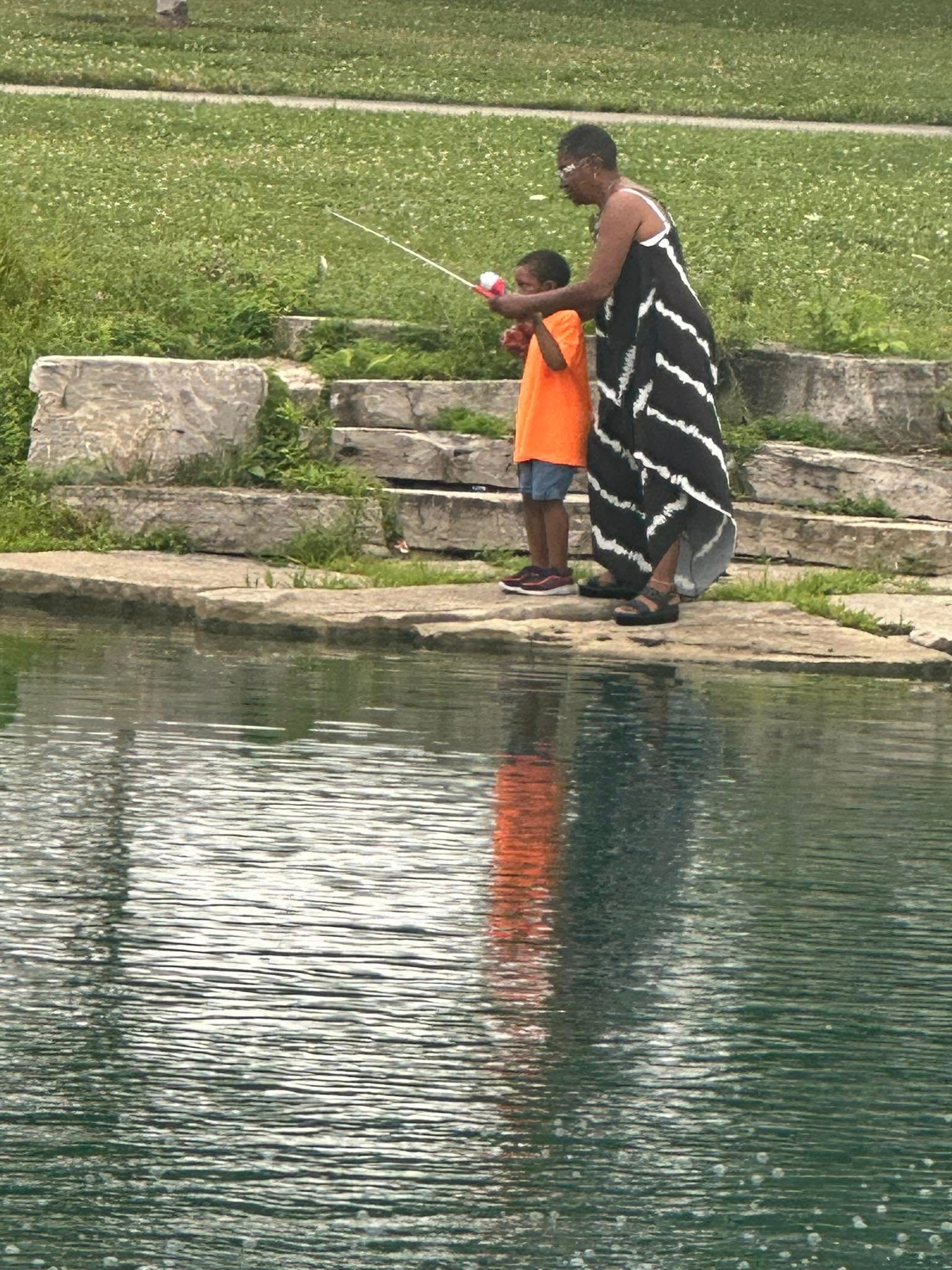 Woman and child fishing by a pond; woman in black and white dress, child in orange shirt.