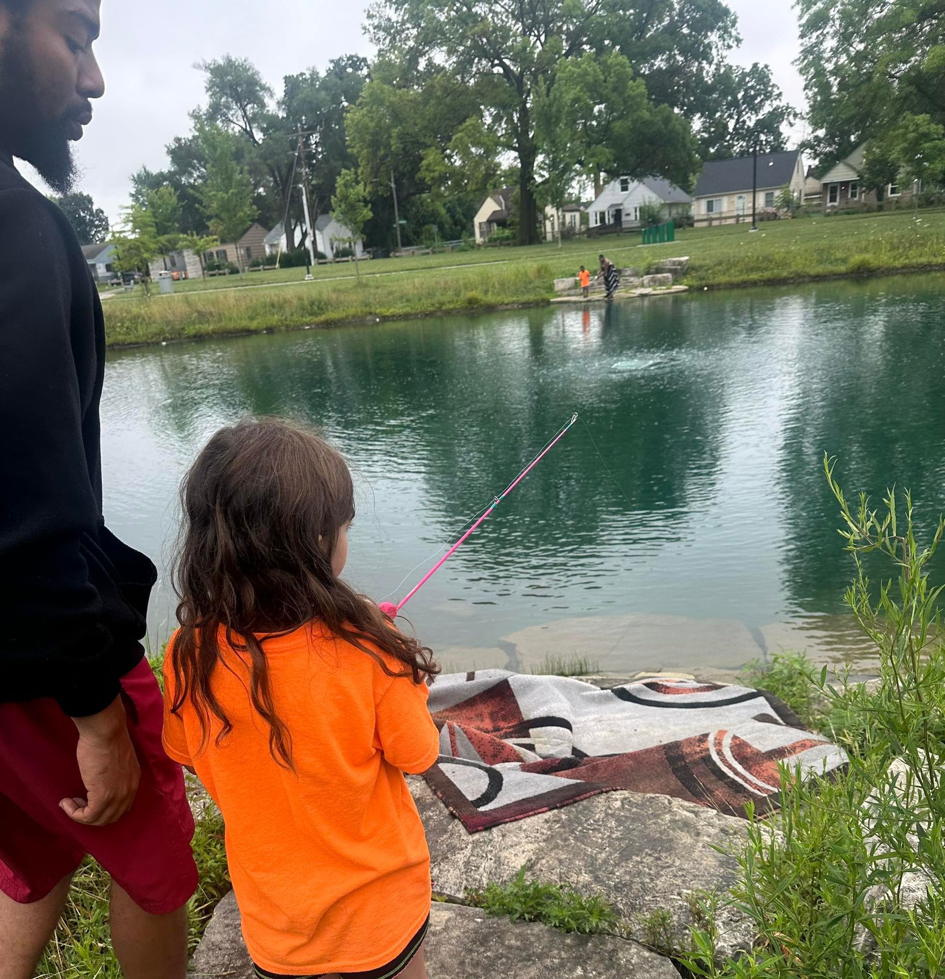 Man and child fishing at a pond. Child holds a pink pole, wearing an orange shirt.
