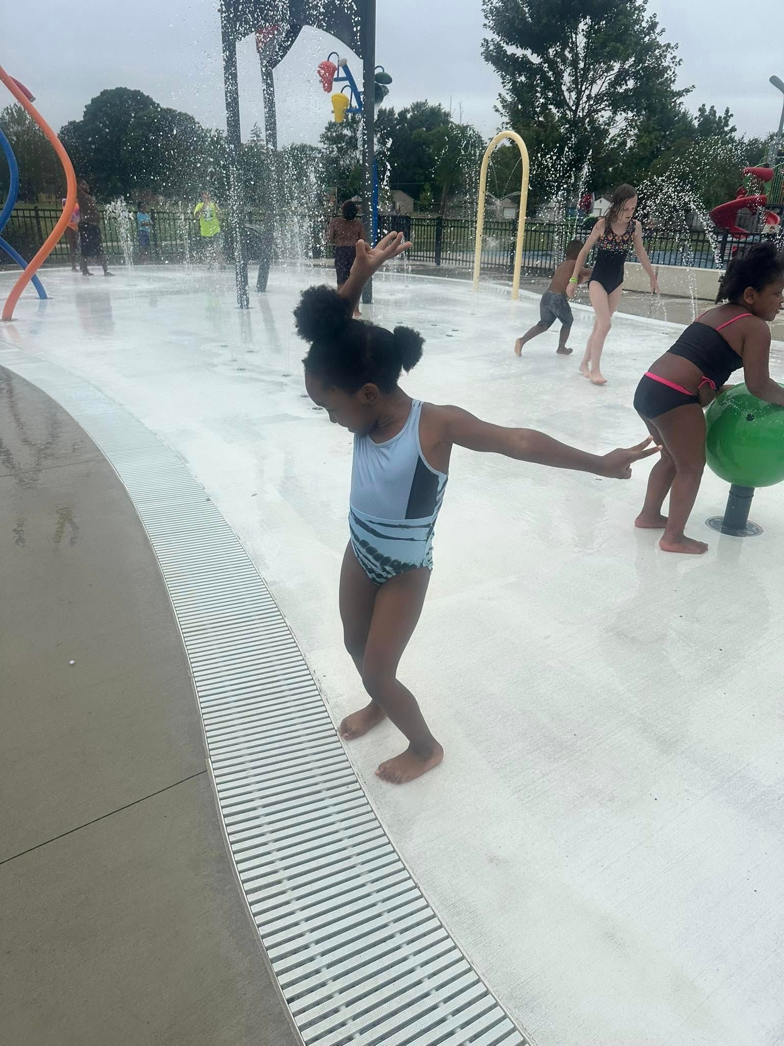 Girl in swimsuit balances on edge of splash pad, arms out. Other children play nearby.