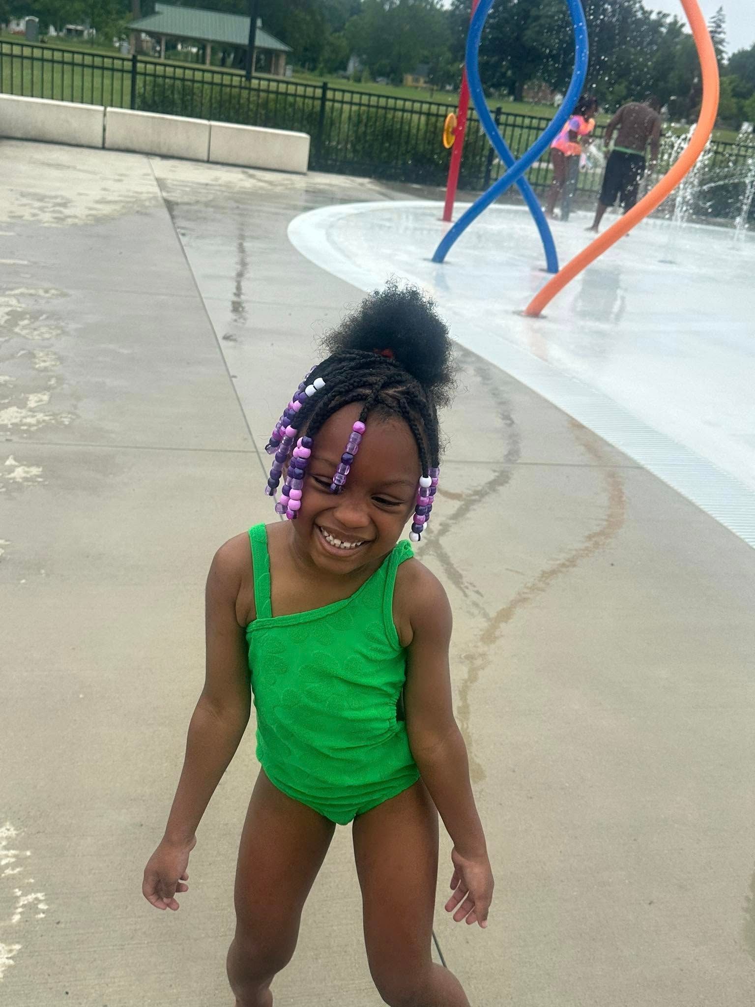 Young girl in green swimsuit smiles at a splash pad. She has beaded braids.