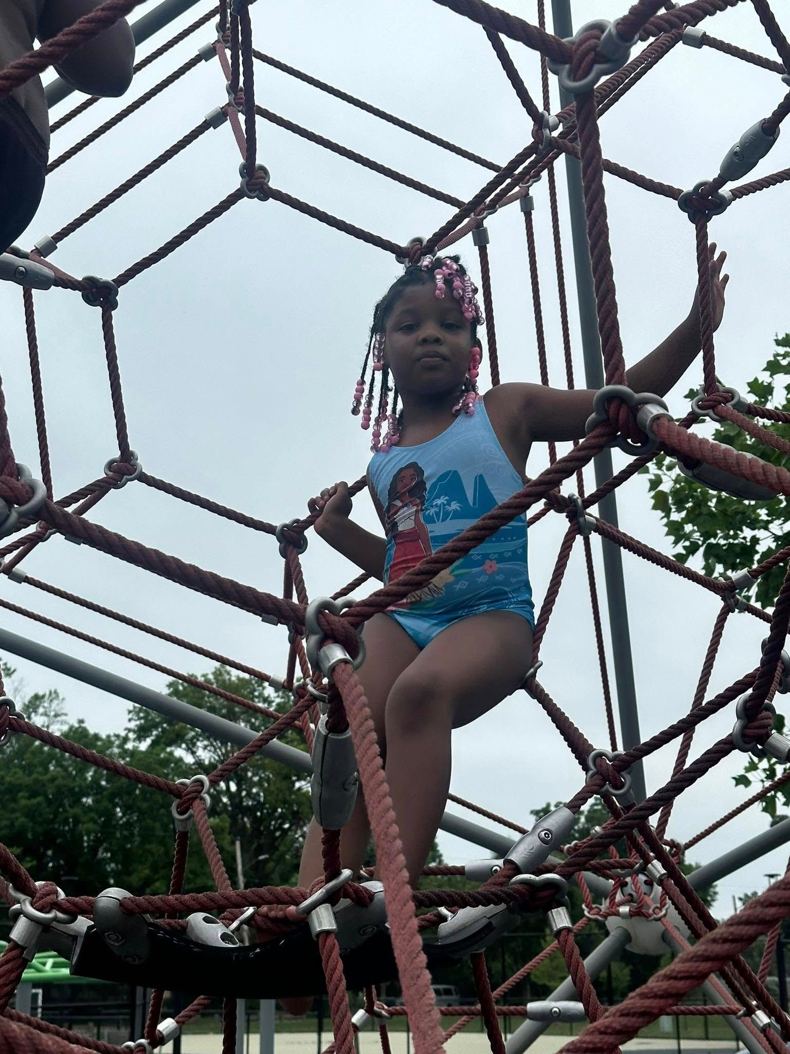 Girl in swimsuit on a playground climbing net, arms out, cloudy sky.