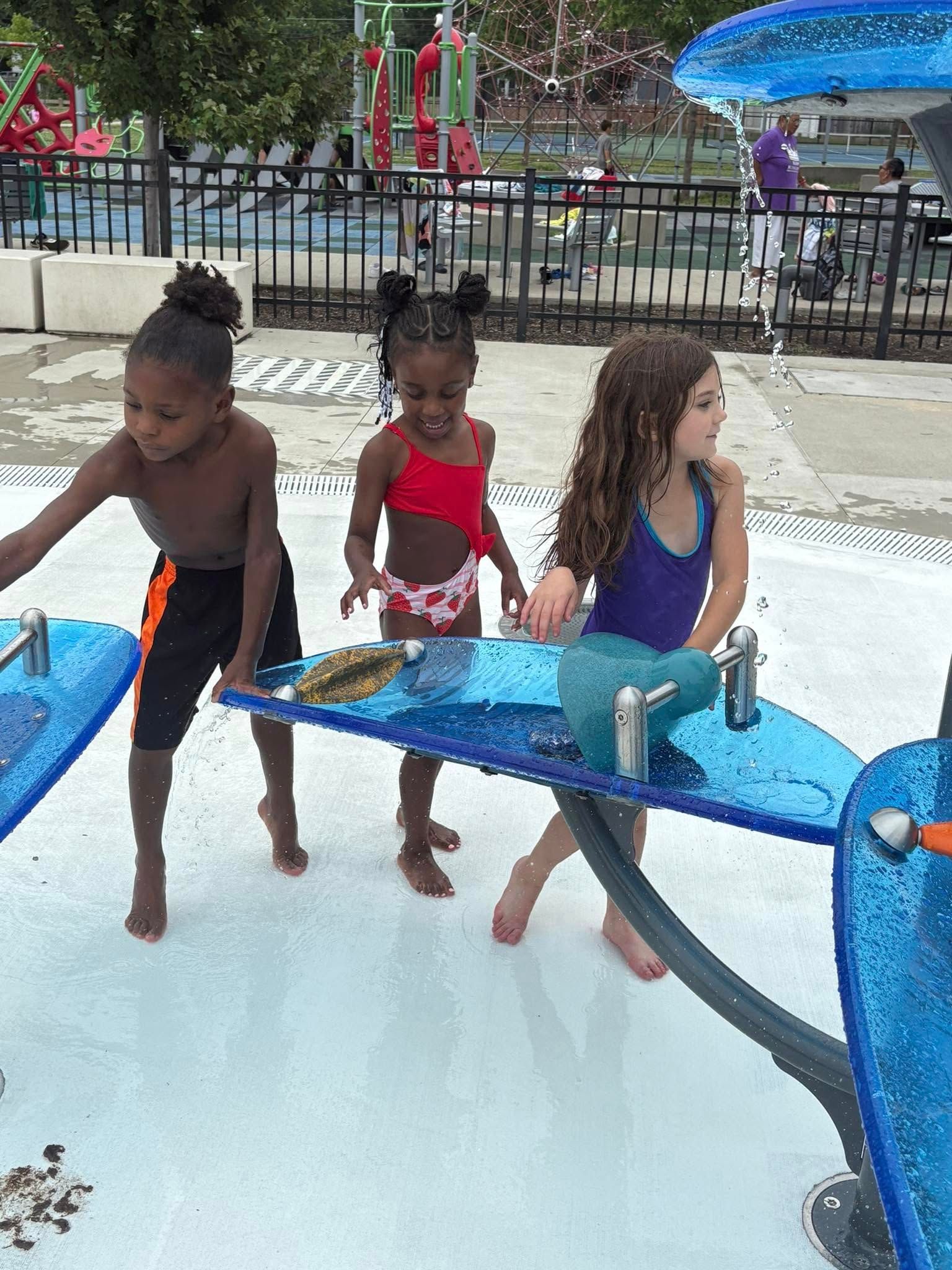 Three children playing in a splash pad; one with curly hair, one in a red swimsuit, and one in black swim trunks.