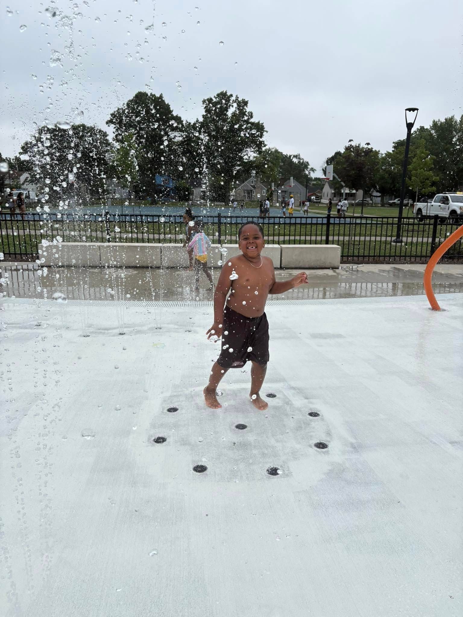 A young man in a splash pad, shirtless, dancing. Water sprays up around him.