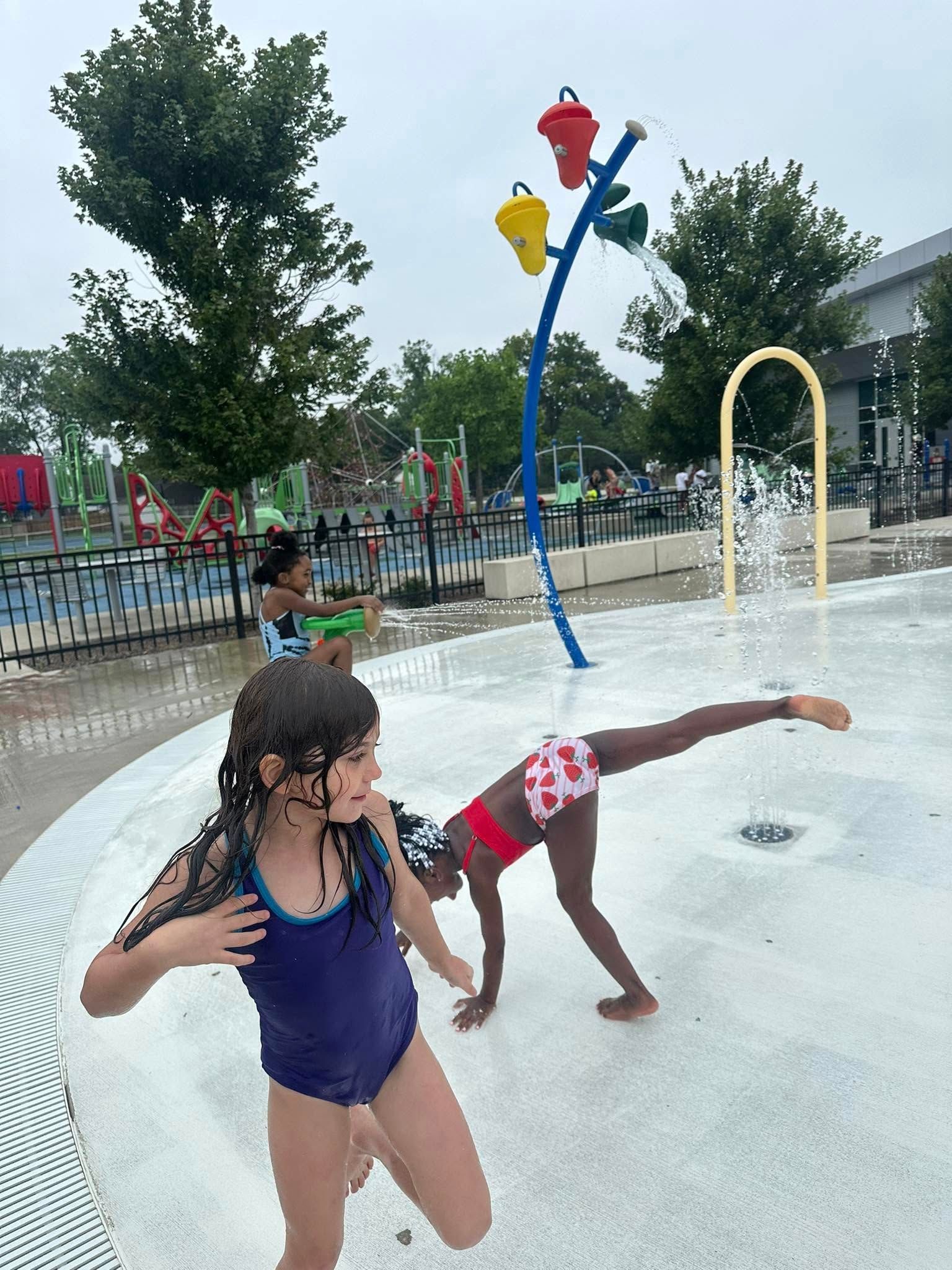 Children playing at a splash pad. One girl cartwheels, another looks surprised, water sprays.