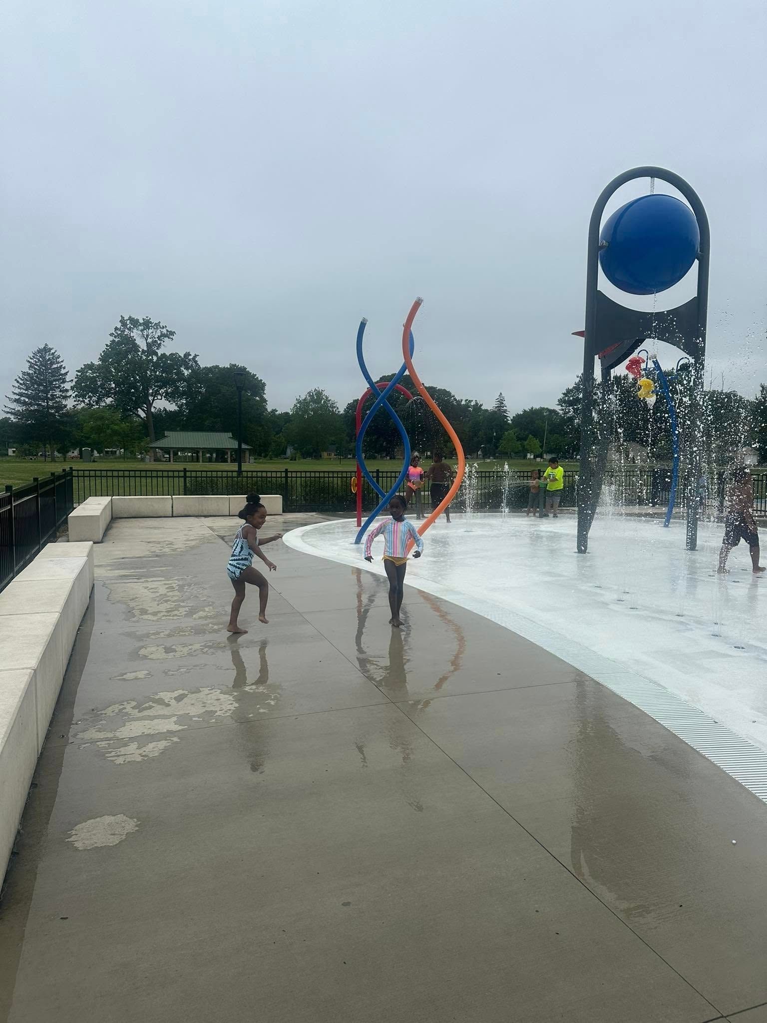 Children playing in a splash pad. Two kids run through water sprays under blue and orange art on a concrete surface.