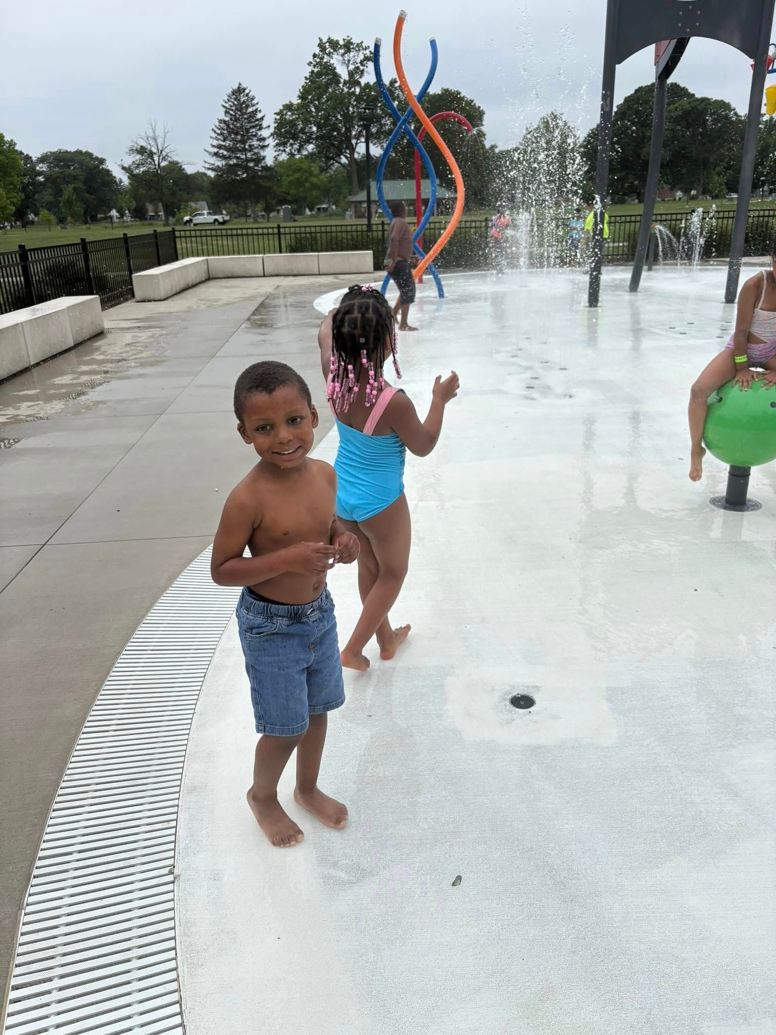 Children playing in a splash pad. Boy in denim shorts smiles; girl in swimsuit. Water sprays.