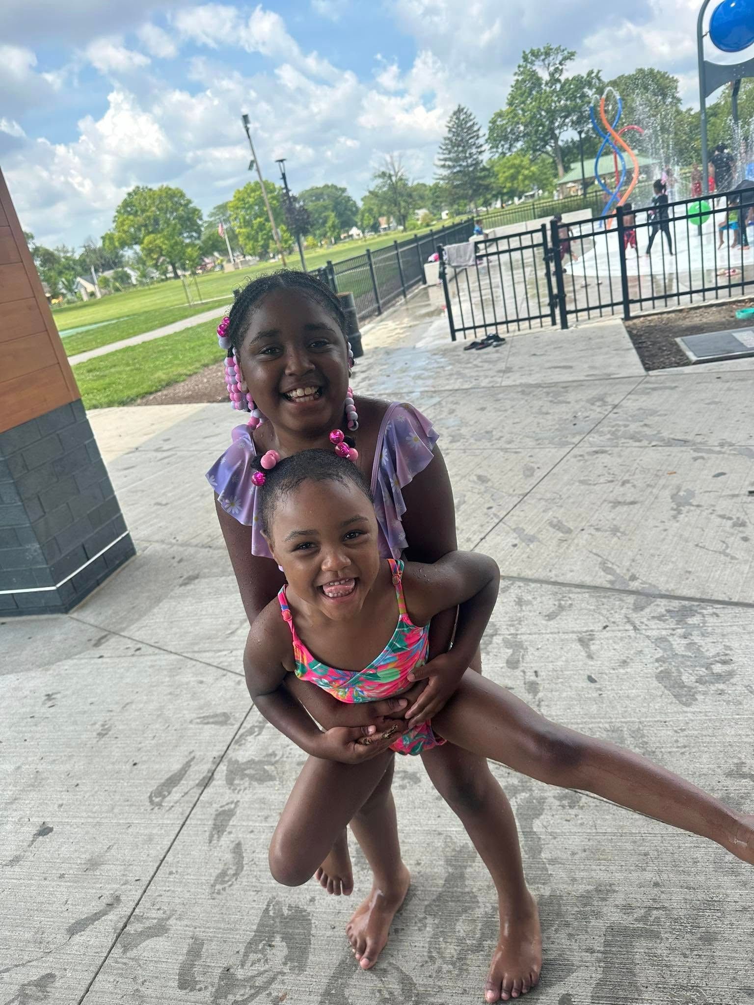 Two young girls smile, one holding the other, at a splash pad. Sunny day, blue sky, green grass.