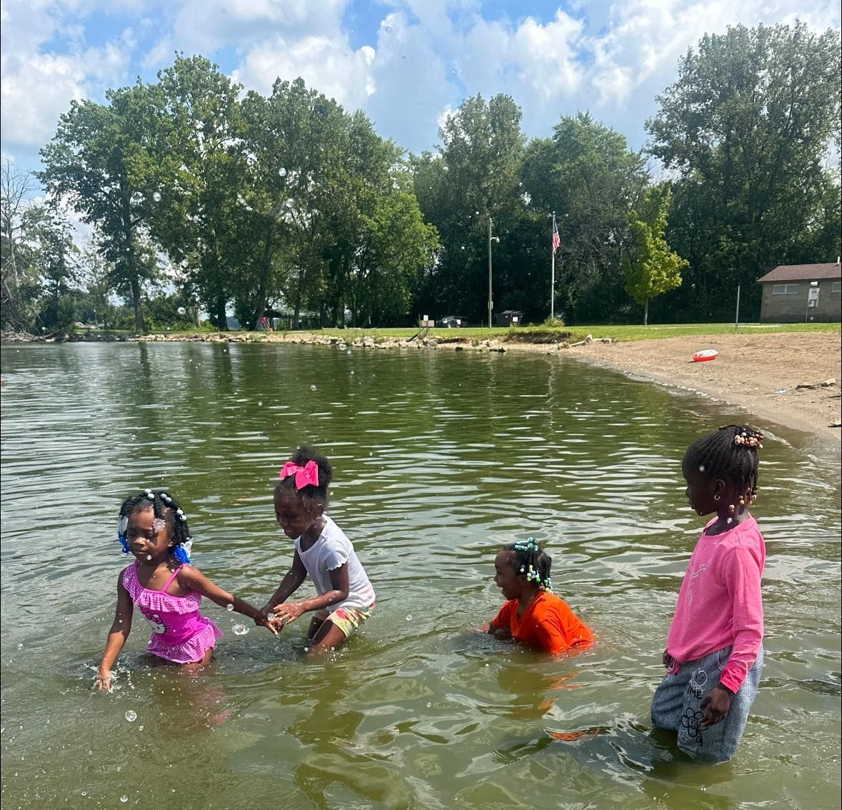 Four young girls playing in a lake near a sandy shore, trees in background.