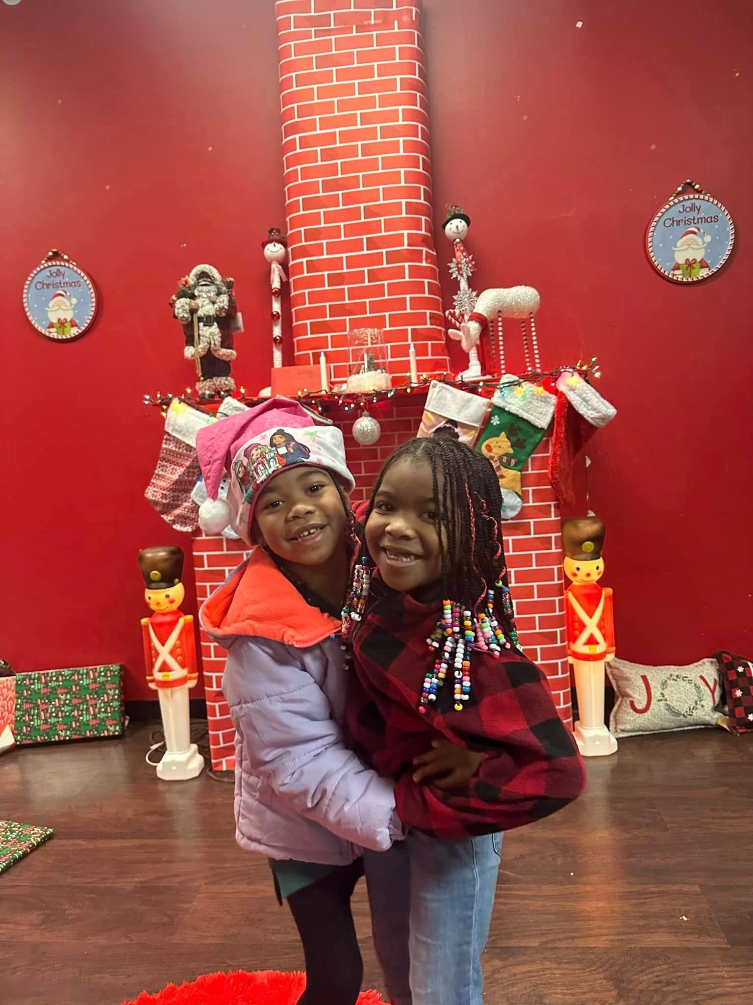 Two girls smiling, hugging in front of a Christmas-themed backdrop with a fireplace and stockings.