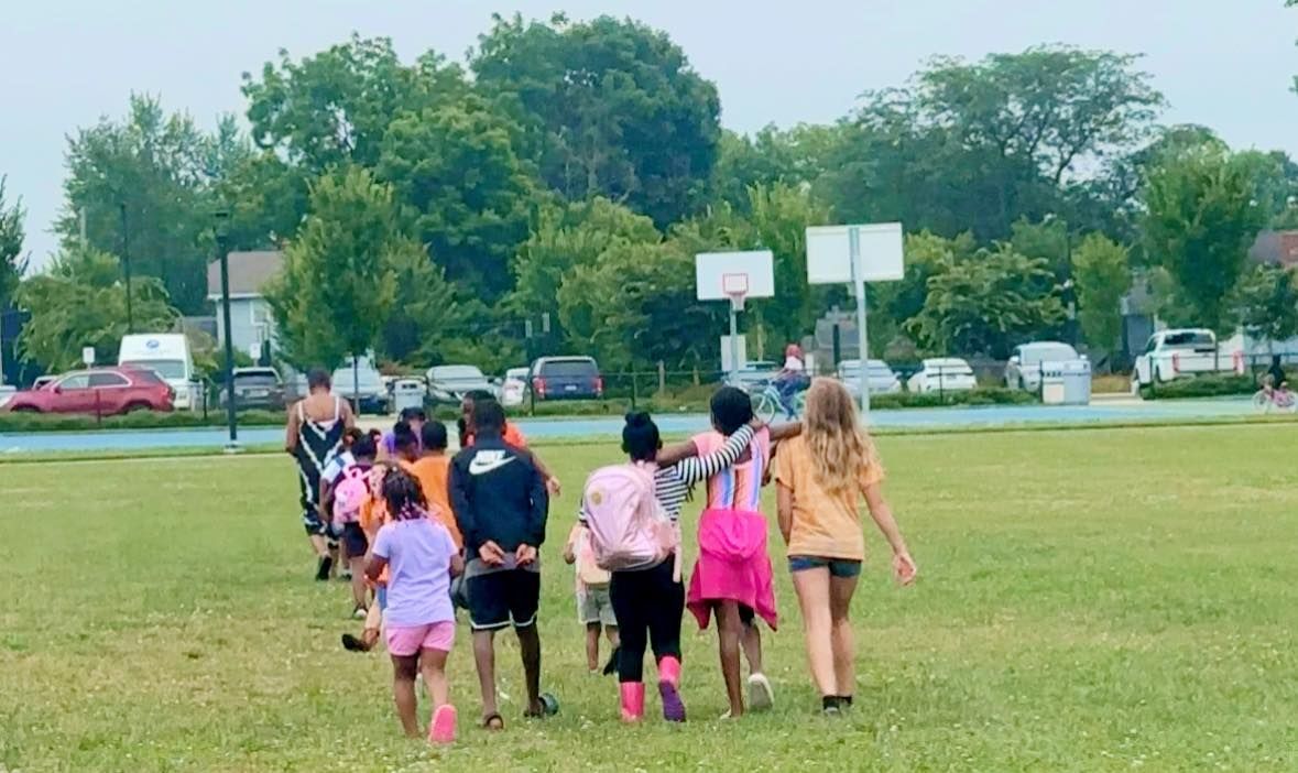 Children walking on a grassy field near a basketball court.