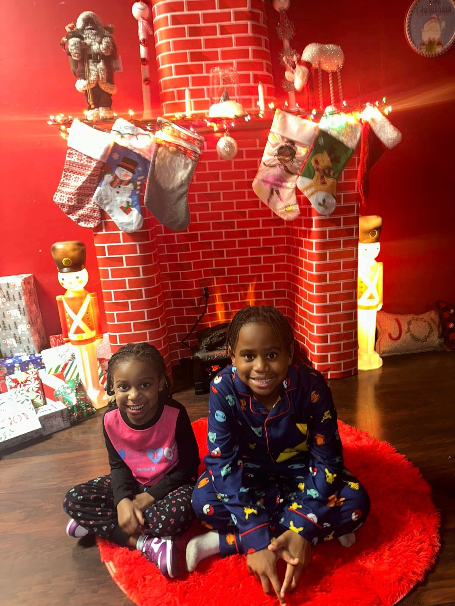 Two young girls in pajamas sit on a red rug in front of a brick fireplace decorated for Christmas.