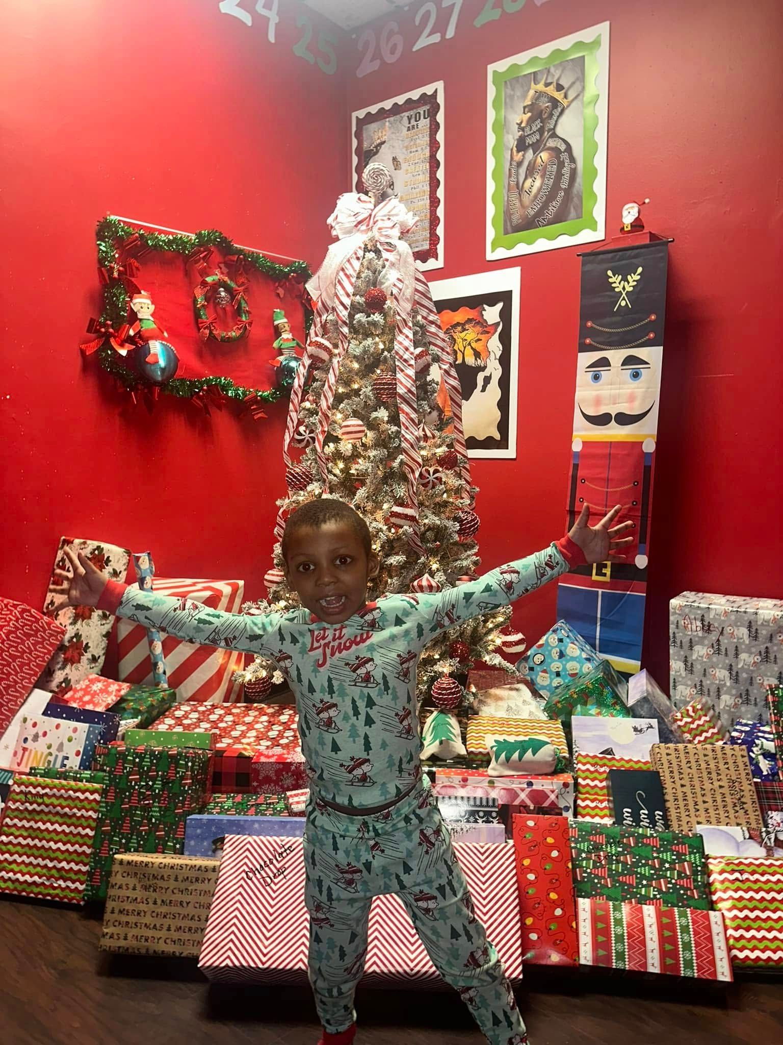 Young child in pajamas with arms outstretched, surrounded by wrapped gifts and Christmas decorations.