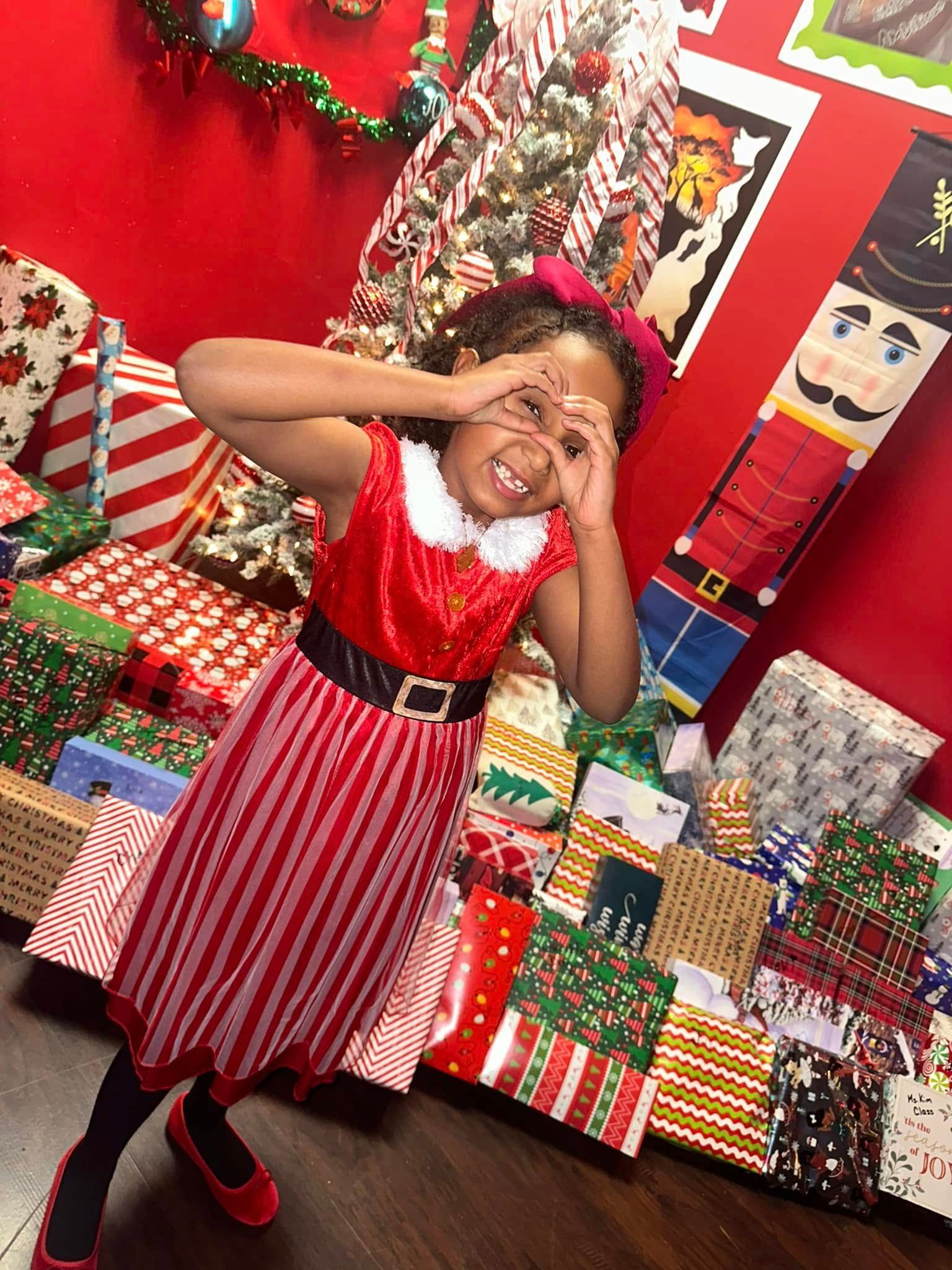 Girl in elf costume making heart shape with hands, smiling near Christmas gifts.