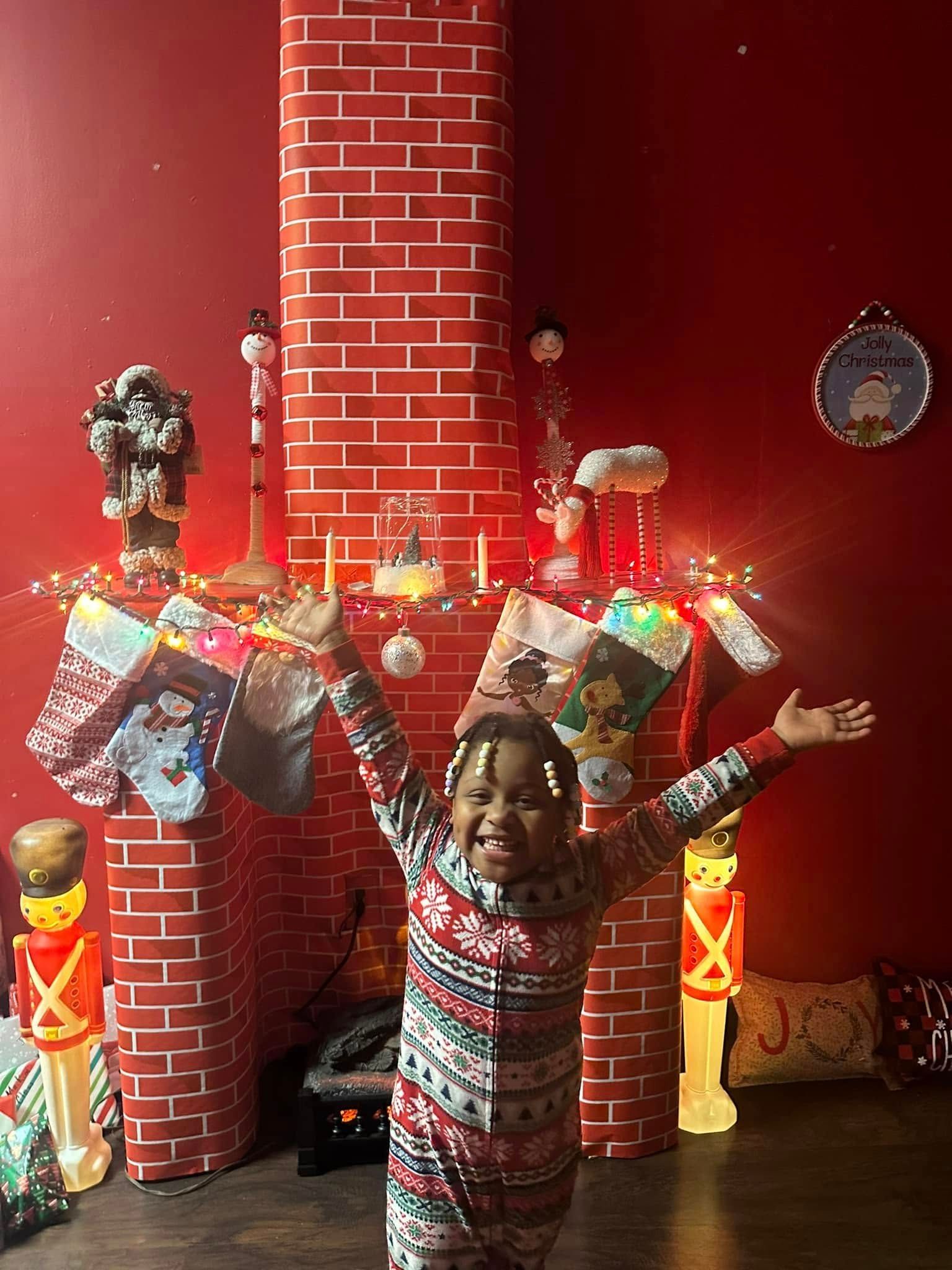Child in Christmas pajamas with arms raised in front of a brick fireplace decorated for Christmas.