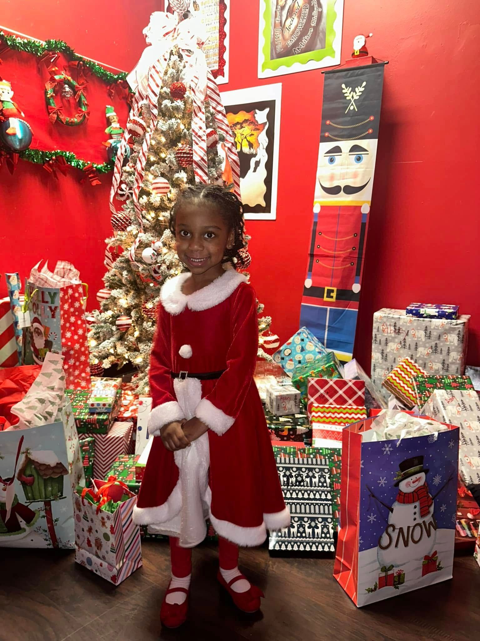 Young girl in a red Santa dress stands in front of a Christmas tree and gifts. Red wall in the background.