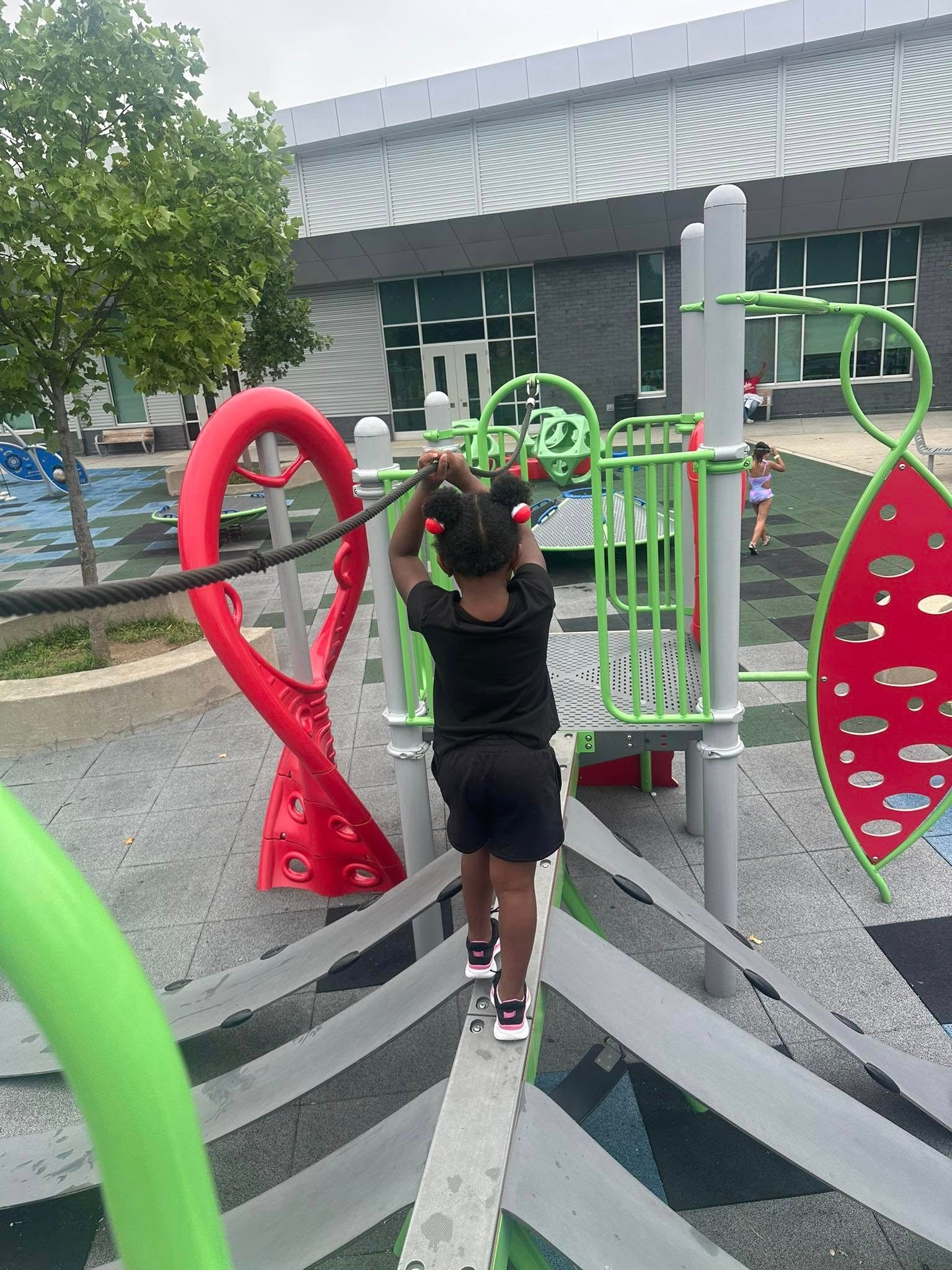 Girl on playground, walking across a balance beam, holding onto a rope, outdoors.