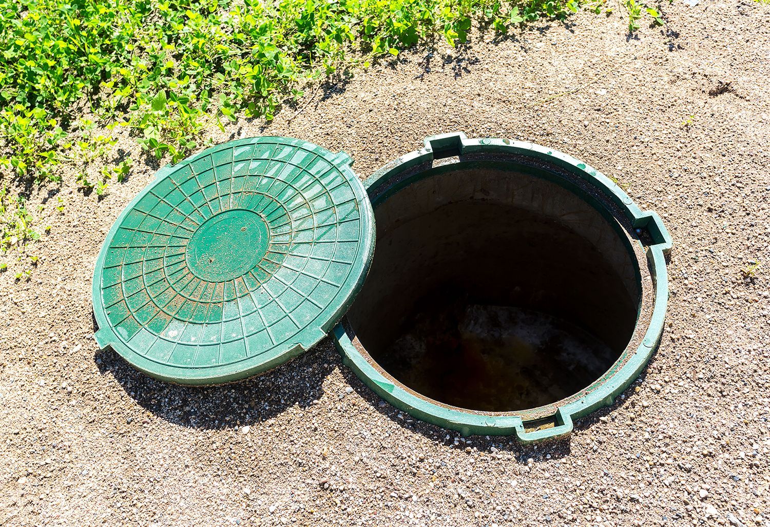 A green manhole cover is open on a dirt ground.