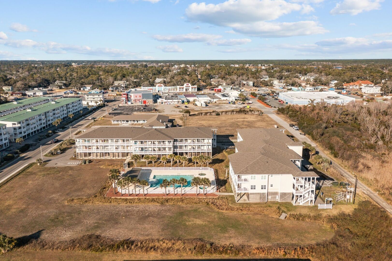 Aerial view of a coastal residential complex with white buildings, pools, and dry grass under a partly cloudy sky
