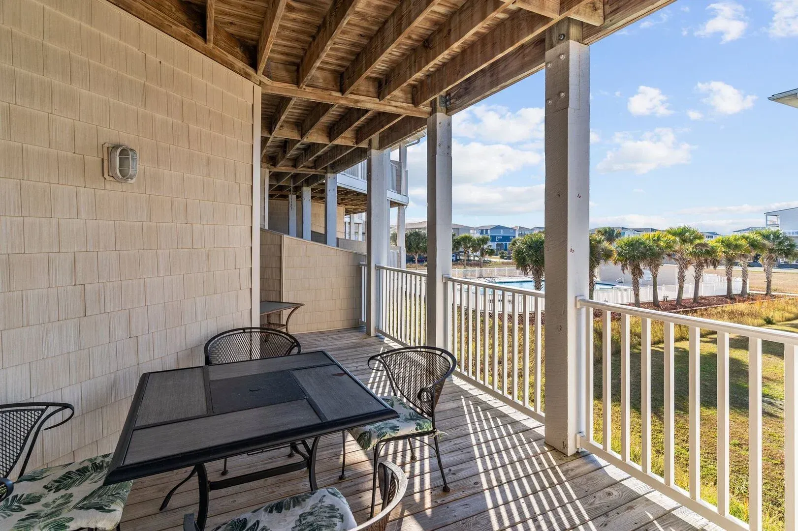 Covered balcony with glass table and chairs, white railing, and a view of palm trees and the beach.