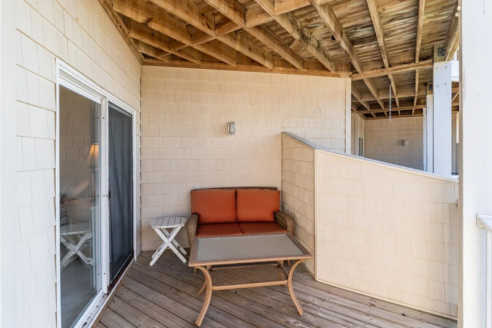 Covered patio with orange loveseat, small table, and exposed ceiling beams above wooden deck.